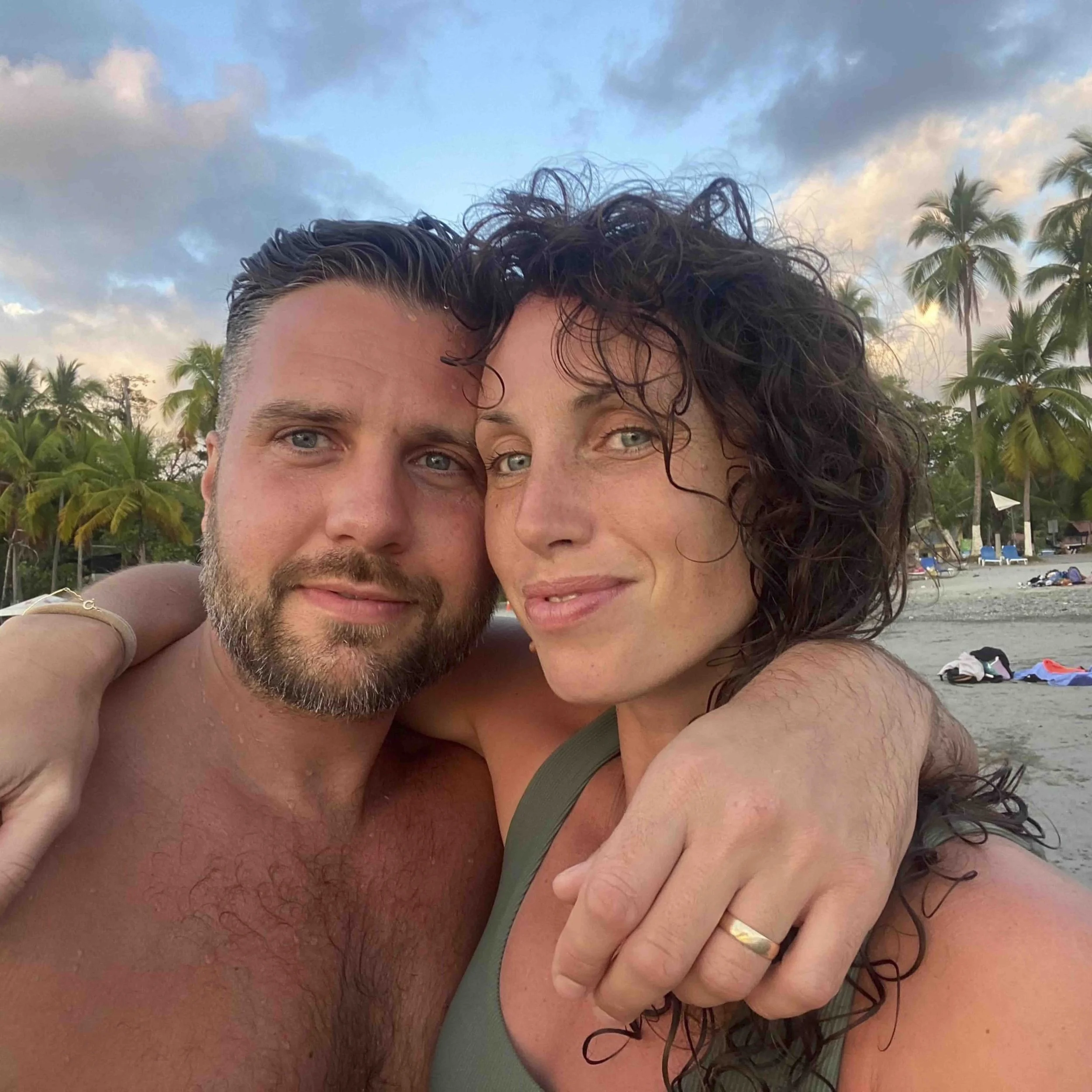 A close-up photo of a man and a woman posing closely together at the beach. The man has blue eyes, light skin, a beard, and short hair. The woman has blue eyes, curly dark hair, and light skin. Palm trees, a cloudy sky, and beach chairs are visible i