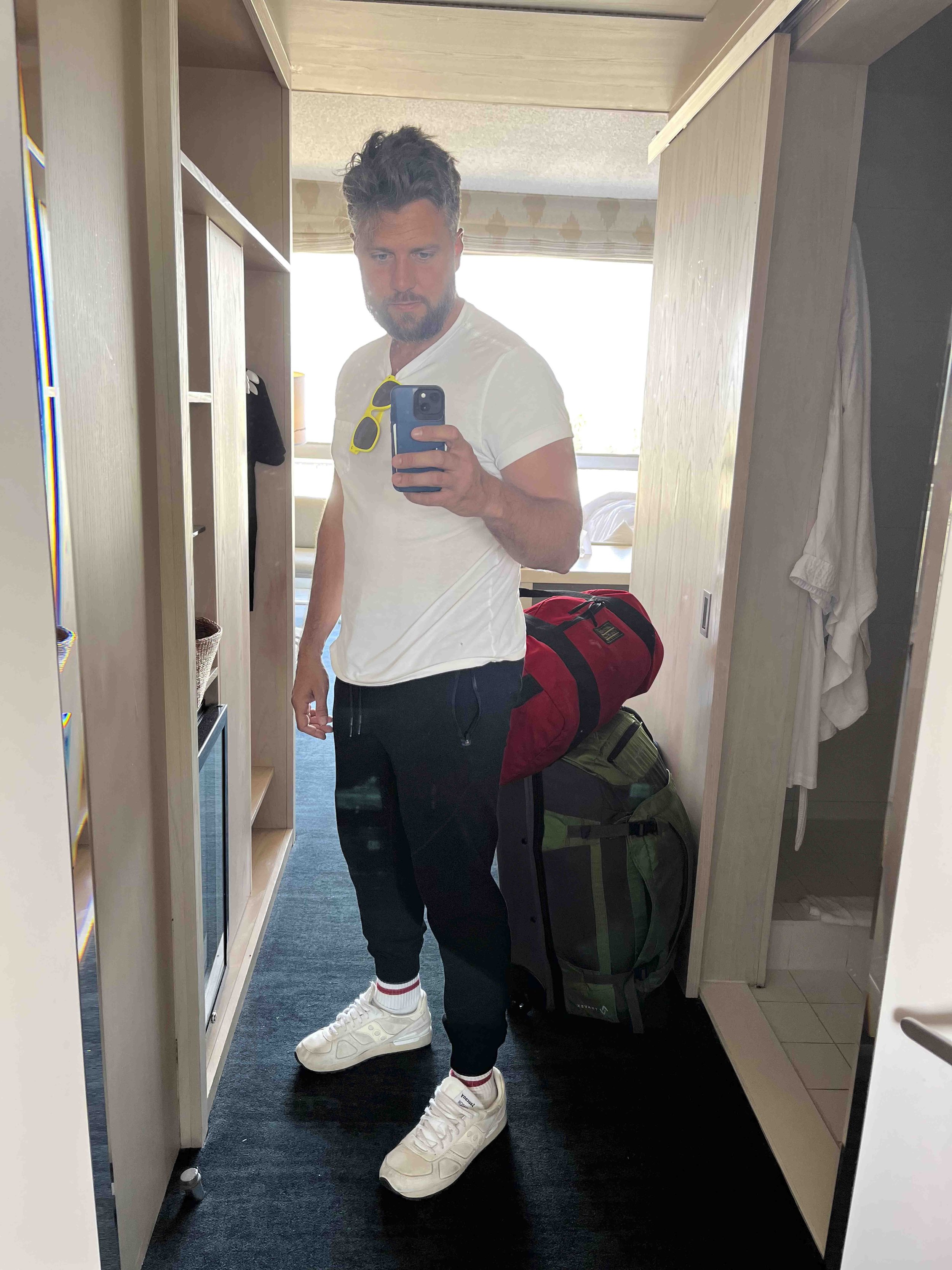 A man taking a mirror selfie in a hotel room. He is wearing a white t-shirt, black pants, white sneakers, and has yellow sunglasses hanging on his shirt. There are two travel backpacks on the floor, and a bed, window, bookshelf, and closet in the bac