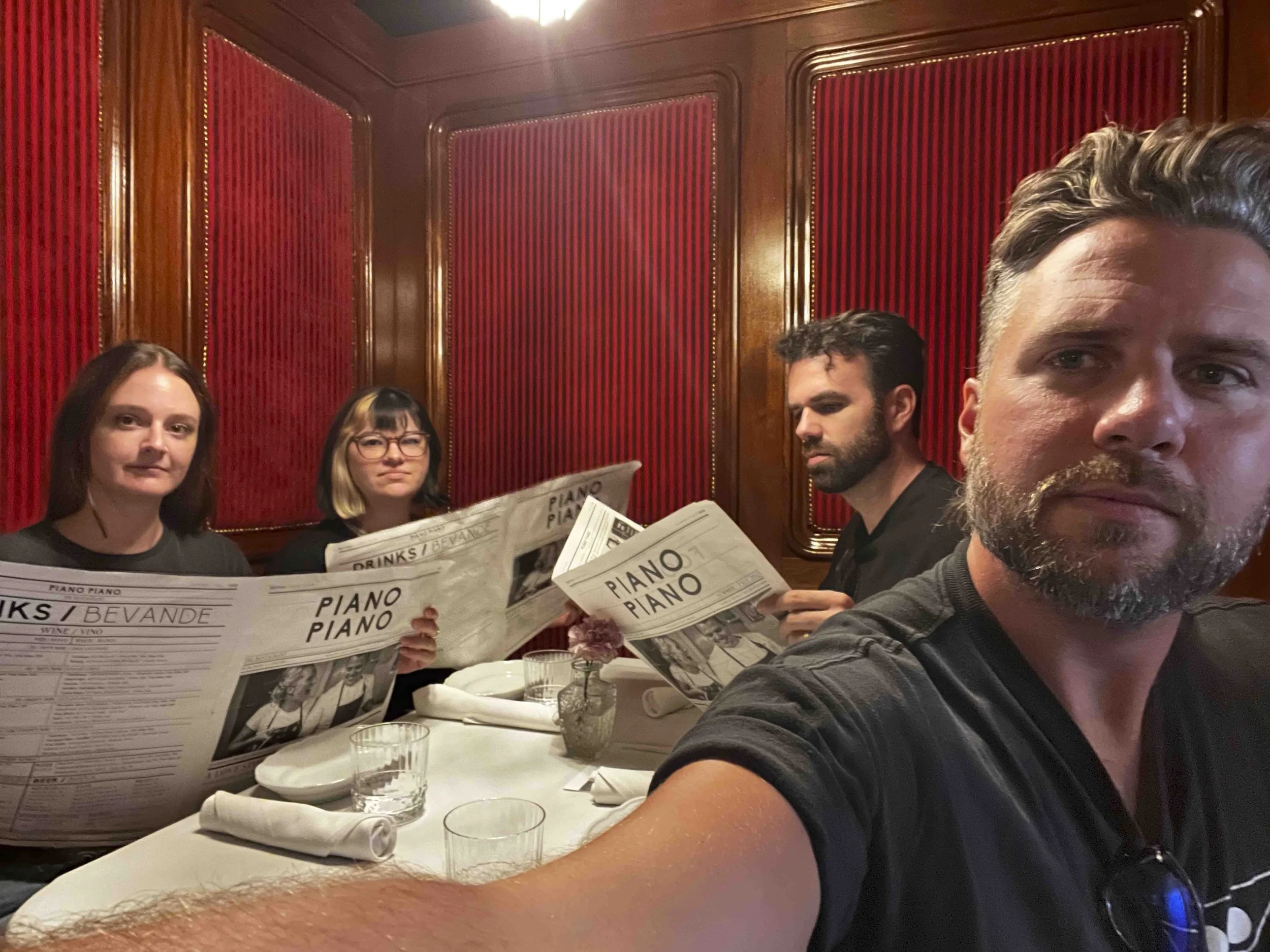 Four people sitting at a restaurant table, reading menus with the word "PIANO" on them, in a room with red striped wall panels and wooden trim, with visible glasses and dishes.
