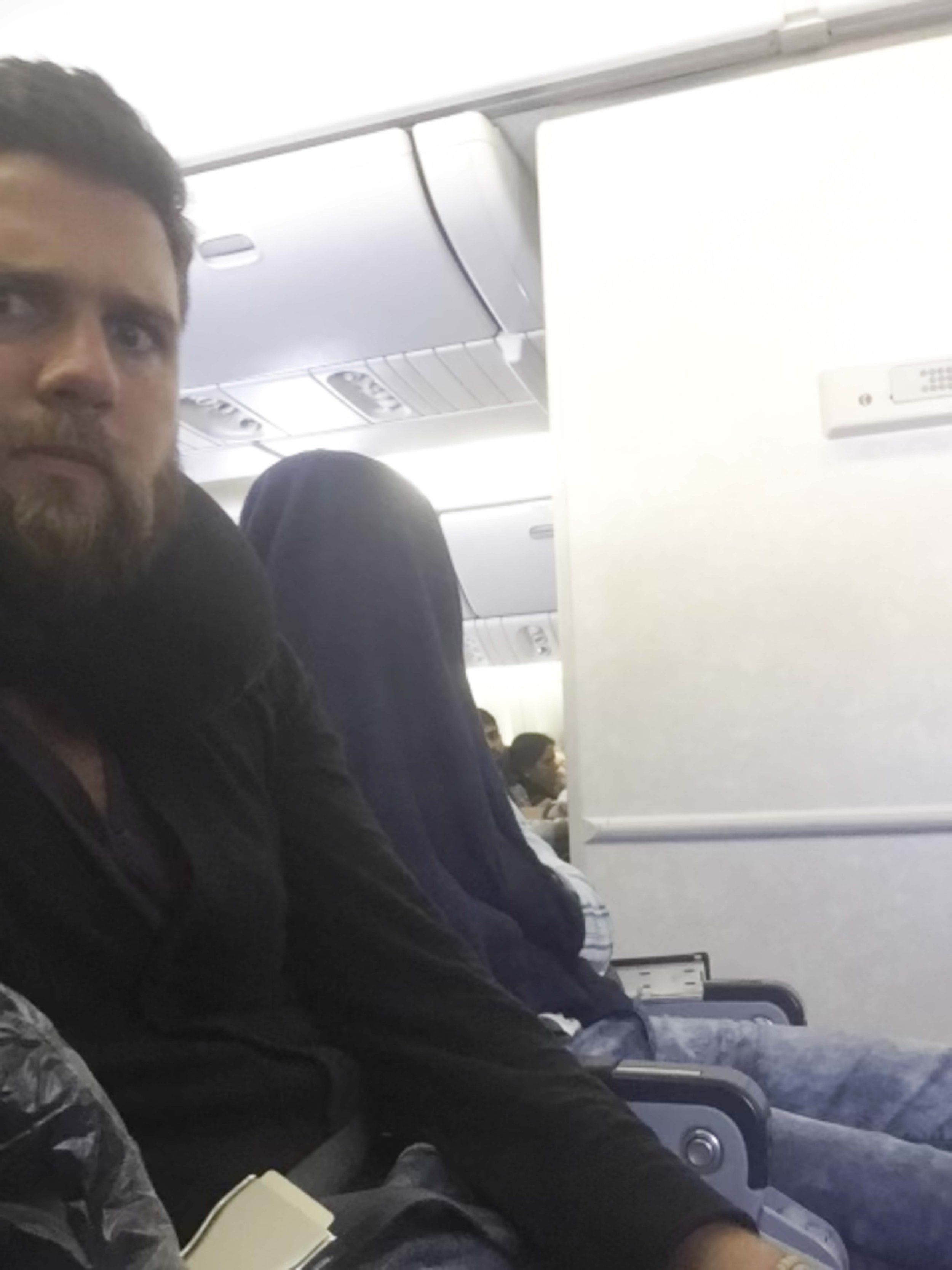 Interior of an airplane cabin with passengers seated, including a man with a beard and dark clothing in the foreground, others visible further back, overhead compartments above, and a white panel on the right side.