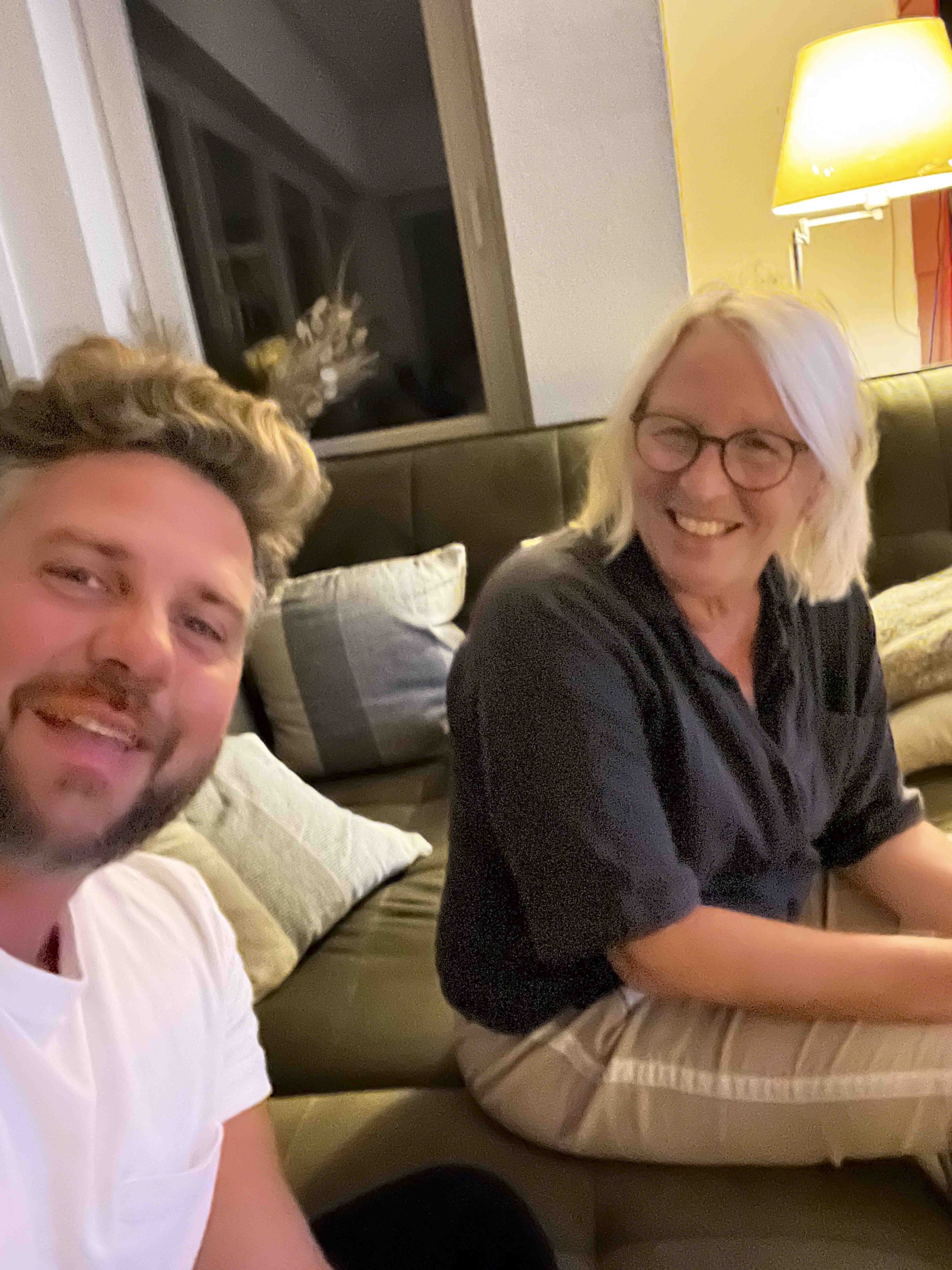 A man with a beard and blond, curly hair taking a selfie with a woman with blond hair and glasses, both smiling, sitting on a brown sofa in a warmly lit living room. A gray and white cat with tentacles is sitting on the windowsill in the background.