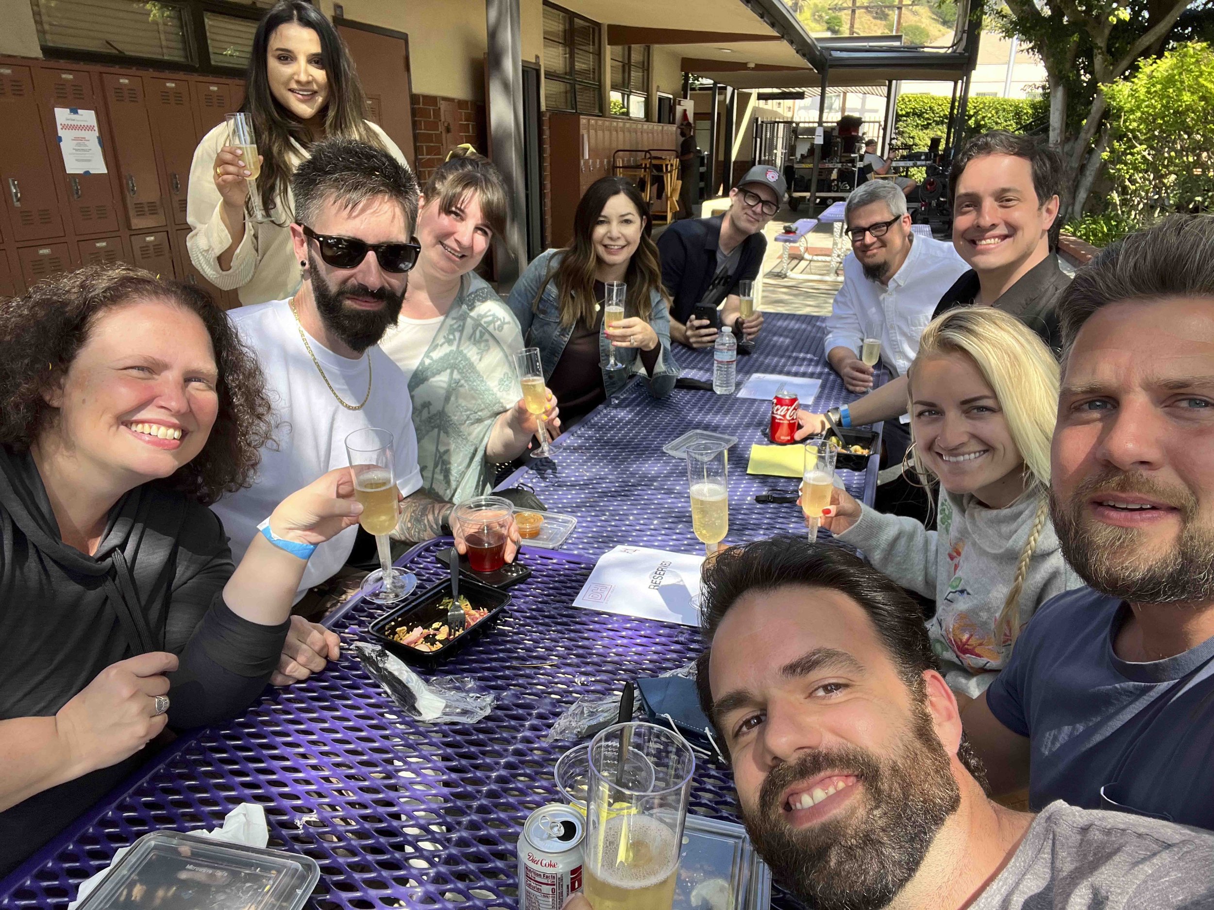 A group of people sitting and standing around a purple outdoor table, smiling and holding drinks at a social gathering. Some have food trays, and one person is using a smartphone. The setting appears to be a patio with lockers, greenery, and a buildi