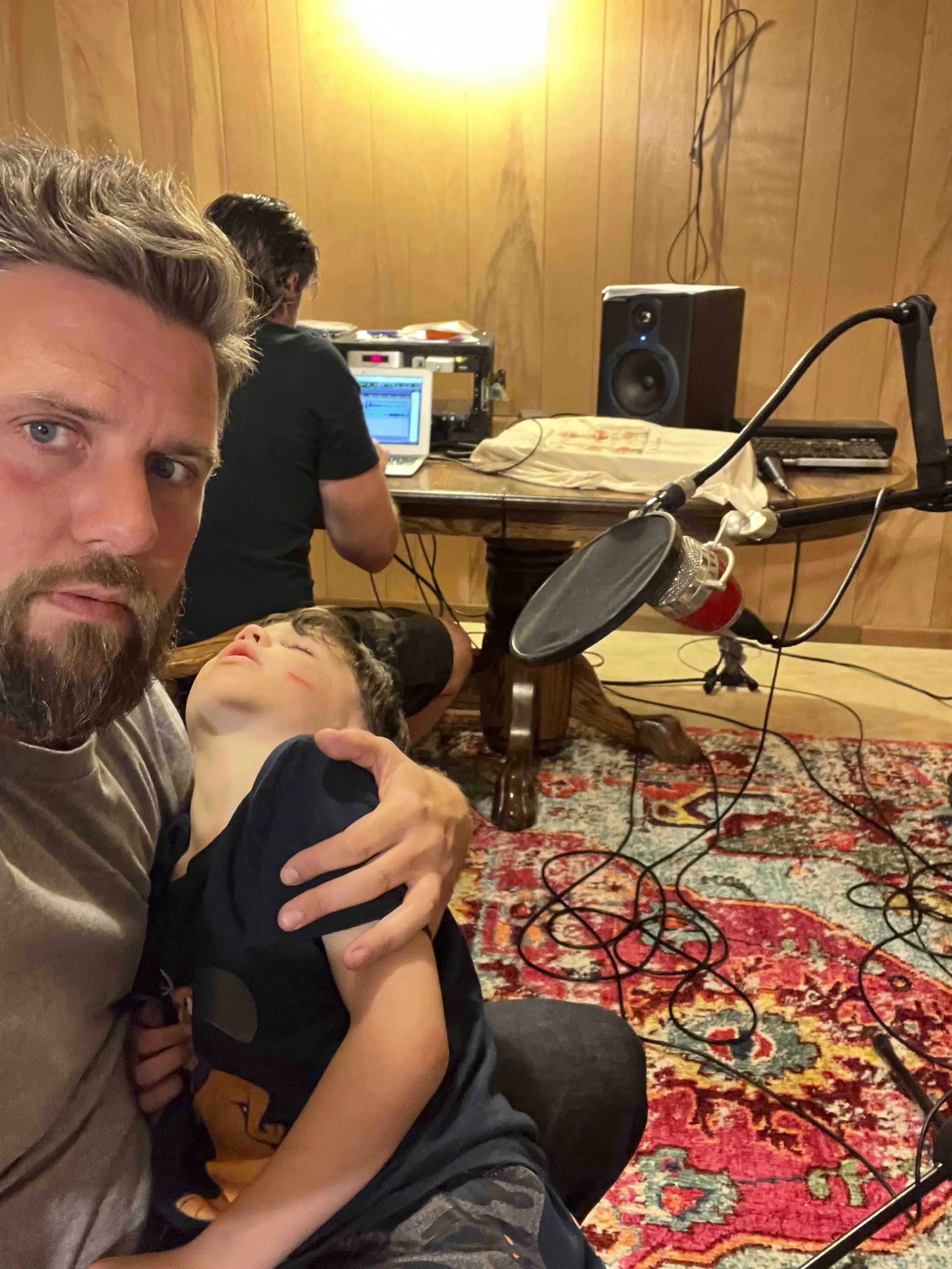 A man with a beard holding a young boy with a bruise on his cheek in a room with wood-paneled walls, a large speaker, a microphone, and a computer setup on a table, with a patterned rug on the floor.