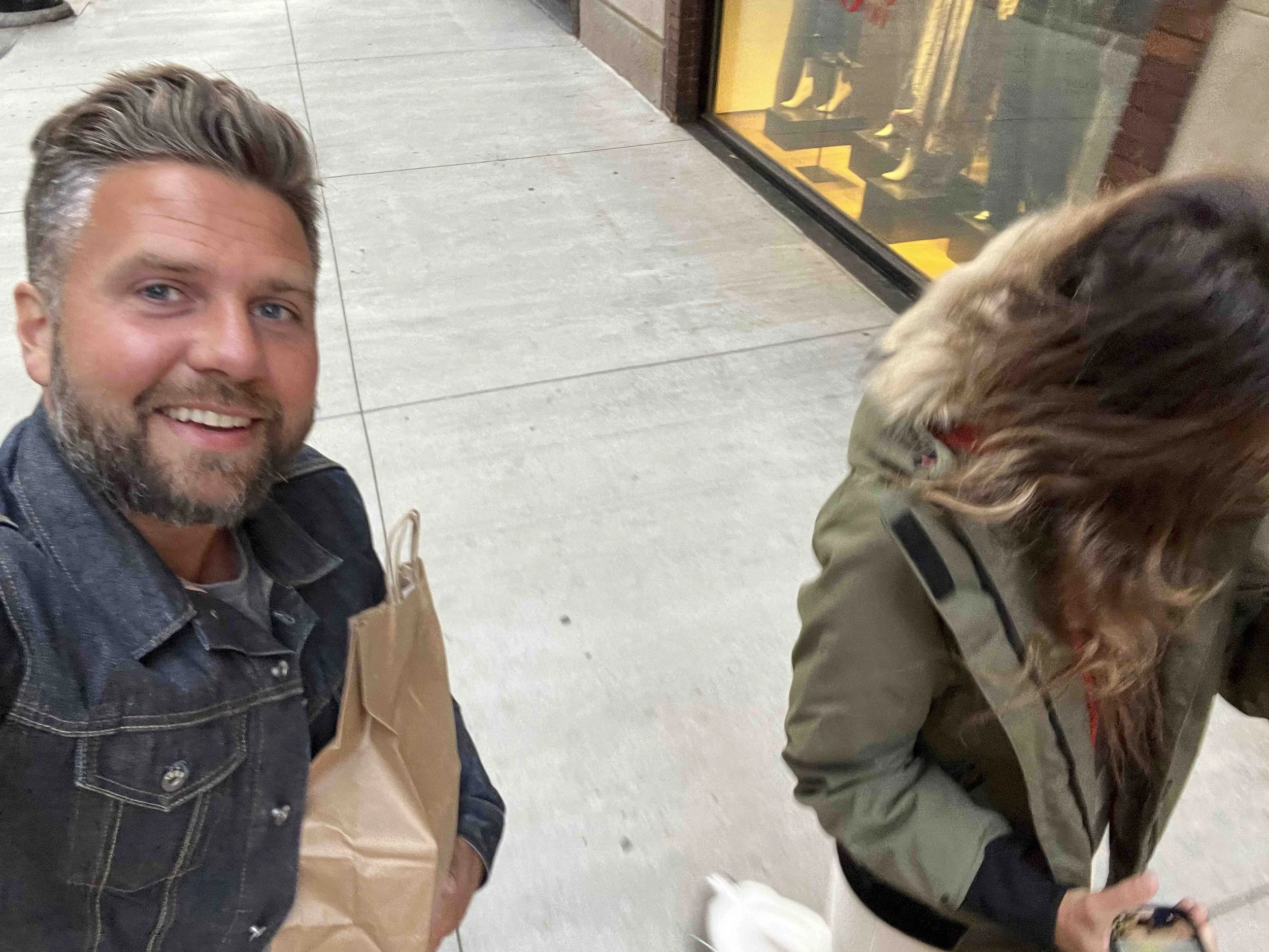A man smiling at the camera with a beard and wearing a dark denim jacket, standing outdoors near a shopping mall entrance. Another person with curly hair, wearing a green jacket, is seated next to him, facing downward. The mall entrance has glass doo