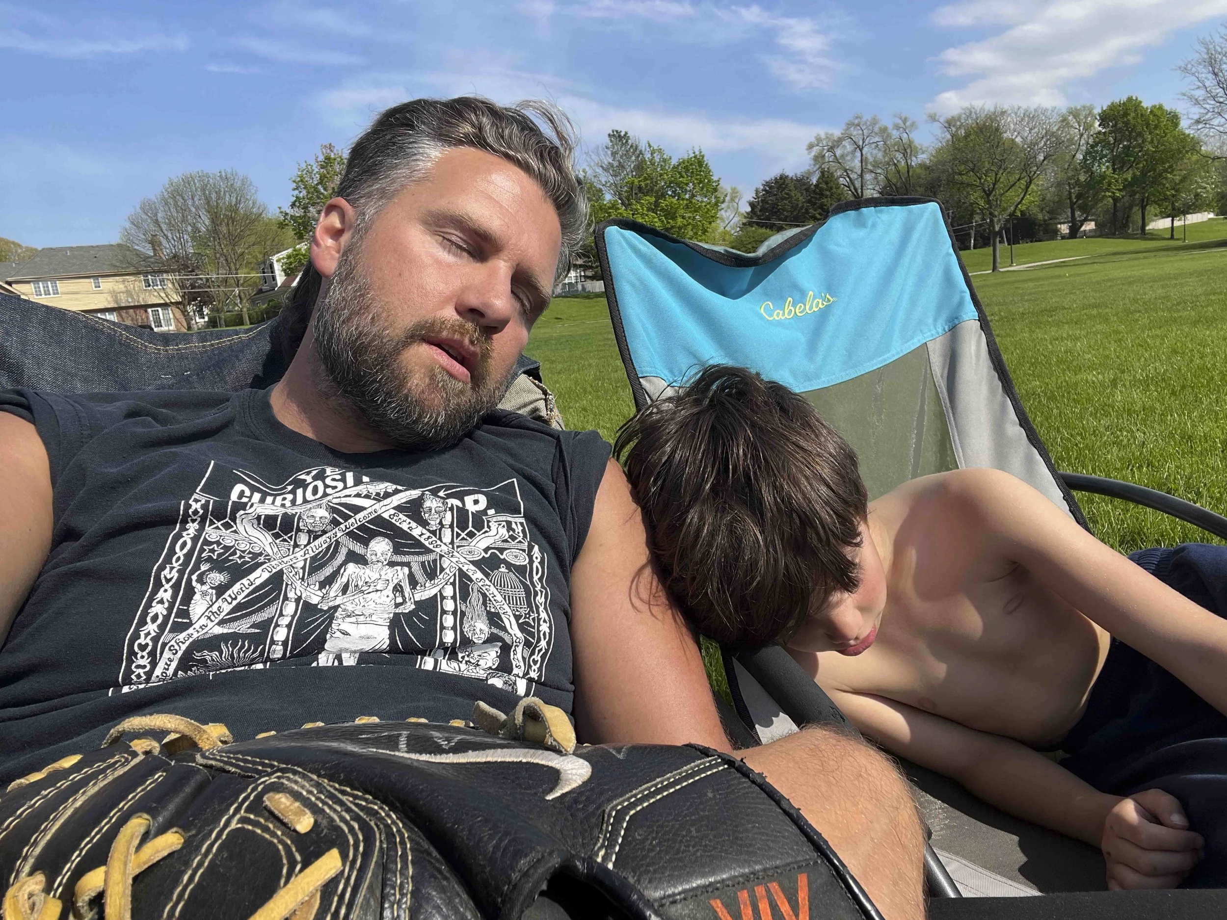 A man with a beard and long hair sleeping outdoors on a sunny day, sitting next to a young boy with short brown hair who appears to be resting or sleeping with his head on the man's arm. They are near a bright blue Cabela's folding chair in a grassy 