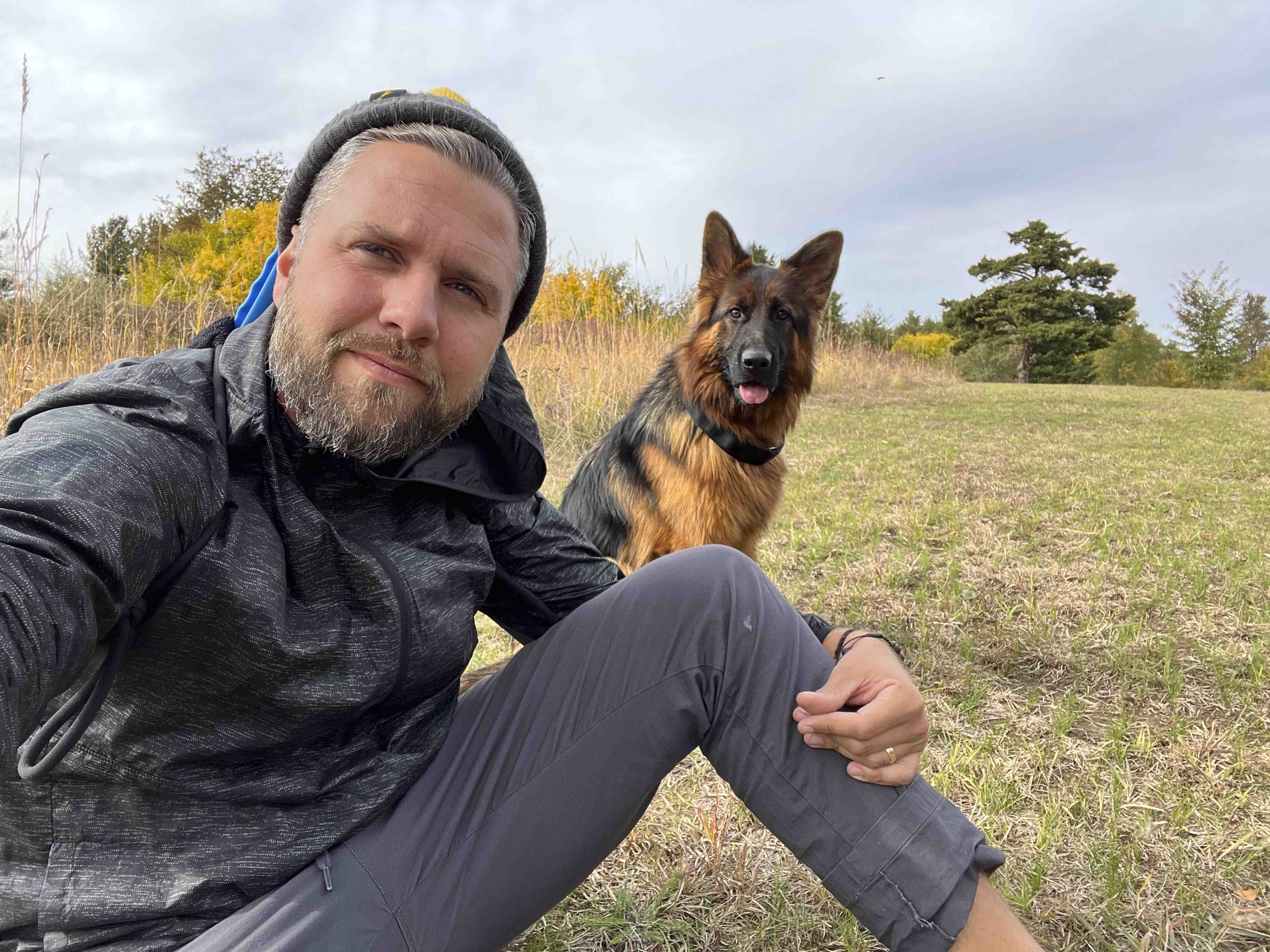 A man with gray hair and beard, wearing a black beanie and jacket, sitting outdoors with a German Shepherd dog on a grassy field with trees and a cloudy sky in the background.