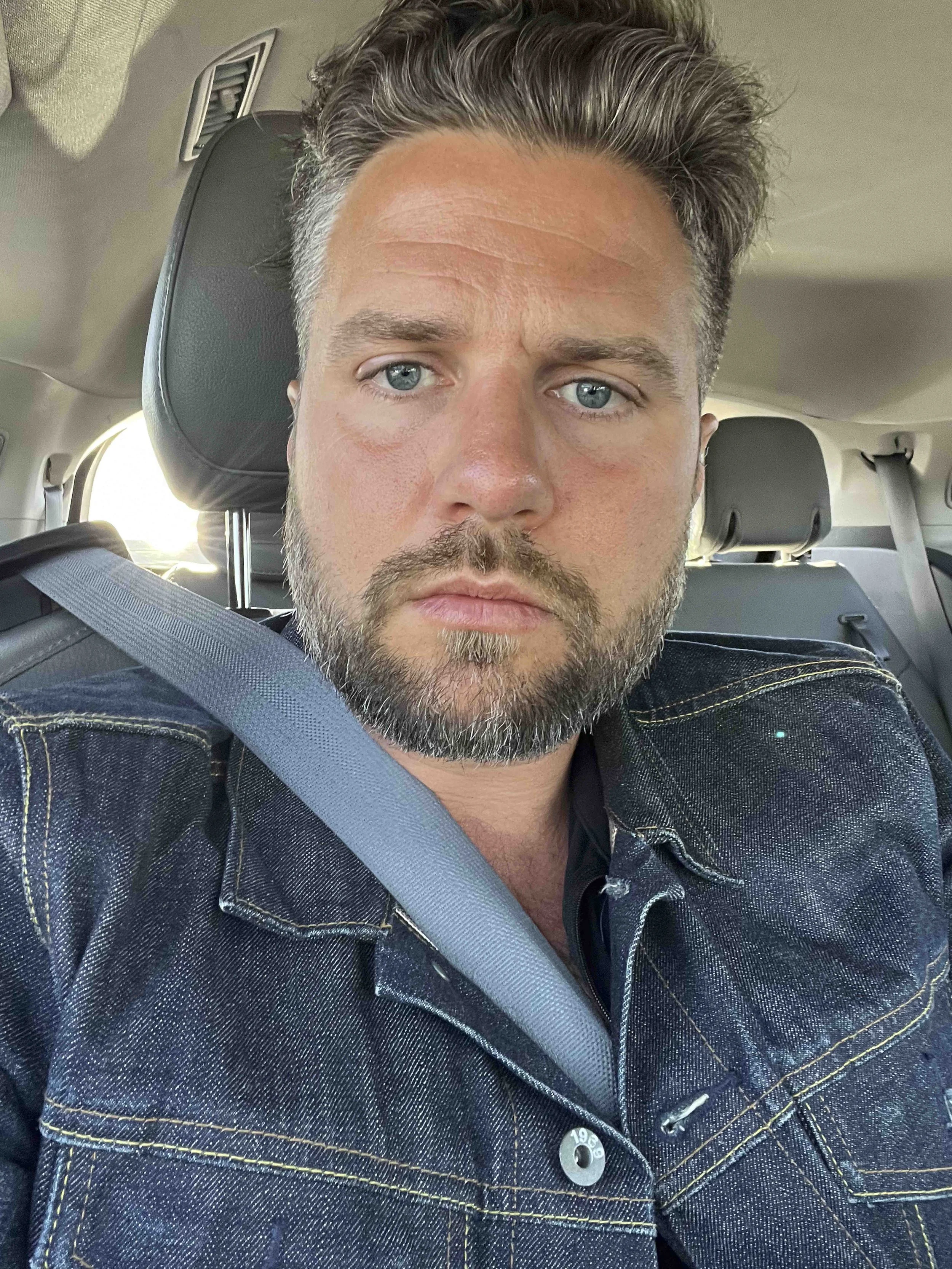 A man with gray hair and a beard sitting in a car, wearing a dark denim jacket and a seatbelt.