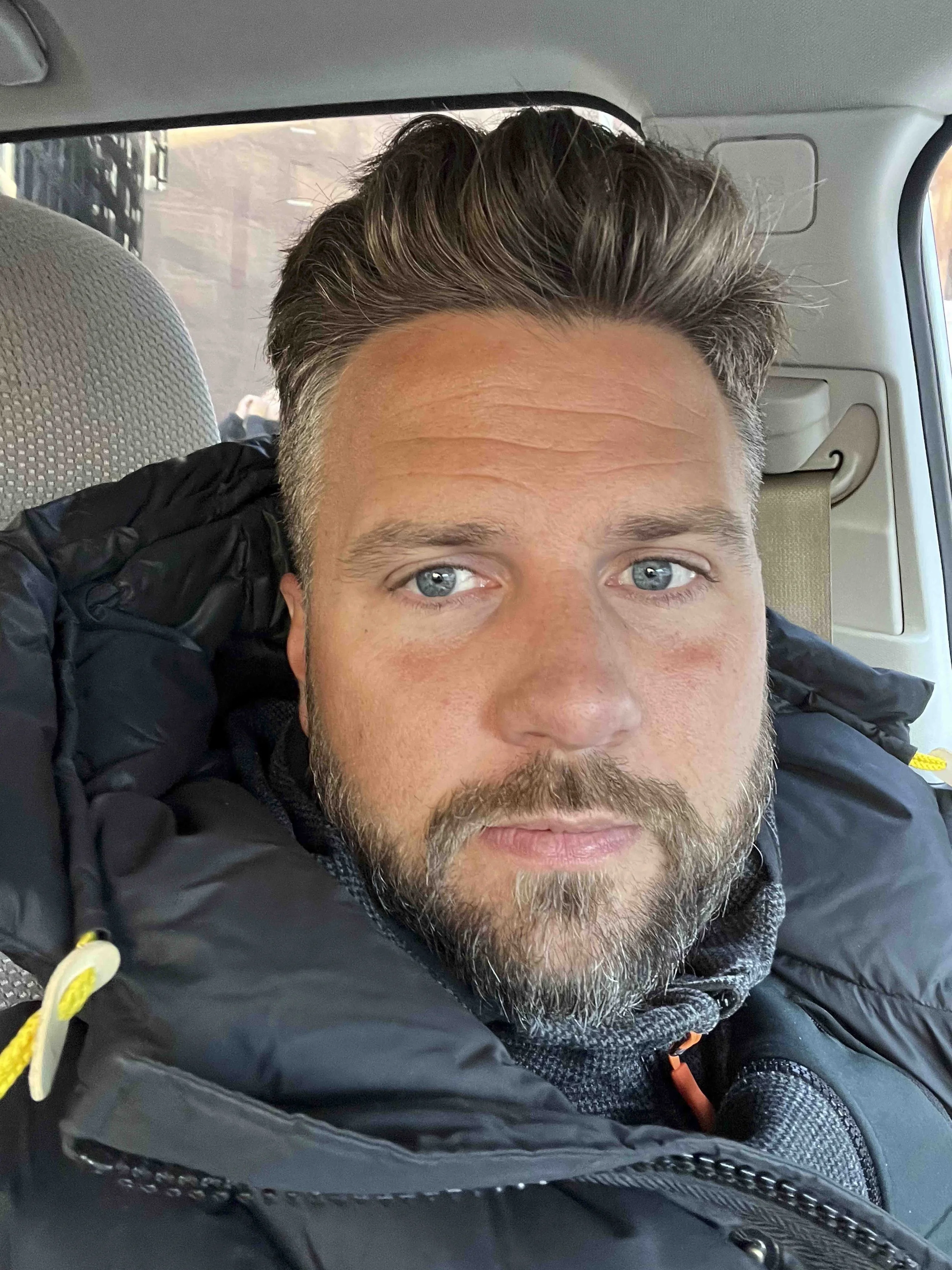 Close-up of a man with short, dark hair and a beard, sitting inside a vehicle, looking directly at the camera.