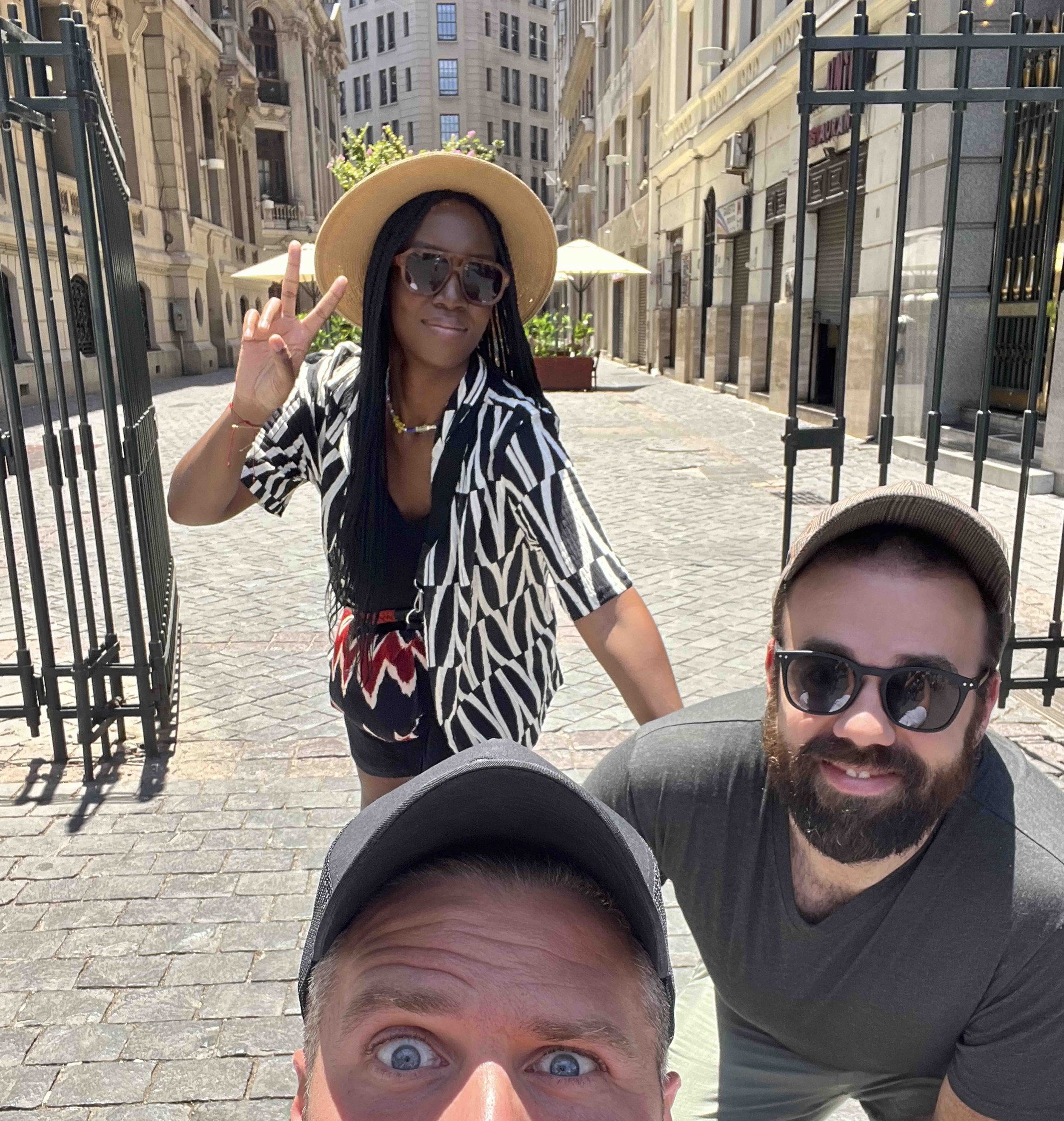 Group of three people taking a selfie on a city street with historic buildings, people, and outdoor umbrellas in the background. One person is wearing a wide-brimmed hat and sunglasses, smiling and making a peace sign.