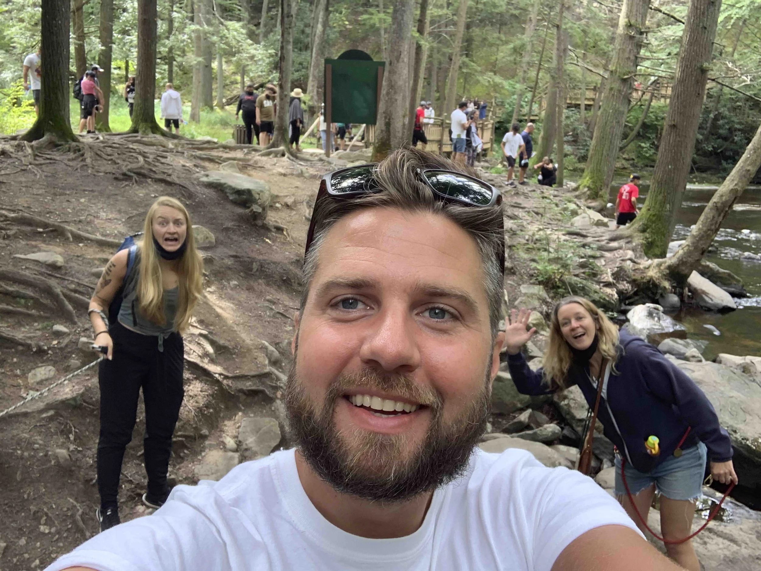 A group of people, including a man with sunglasses on his head and two women, pose for a selfie in a forested area with tall trees and rocky ground.