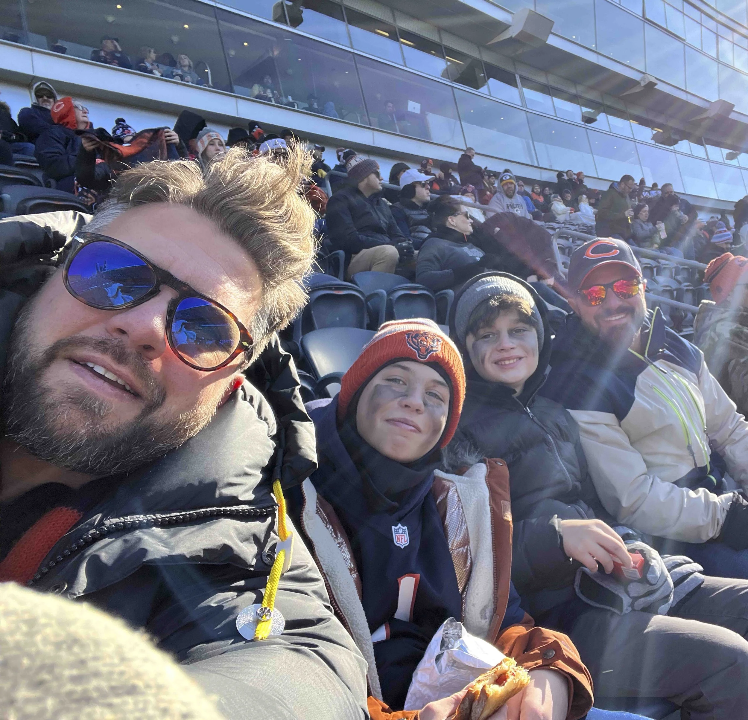 A group of football fans sitting in stadium seats outside a modern glass building, wearing team apparel and enjoying food and beverages.
