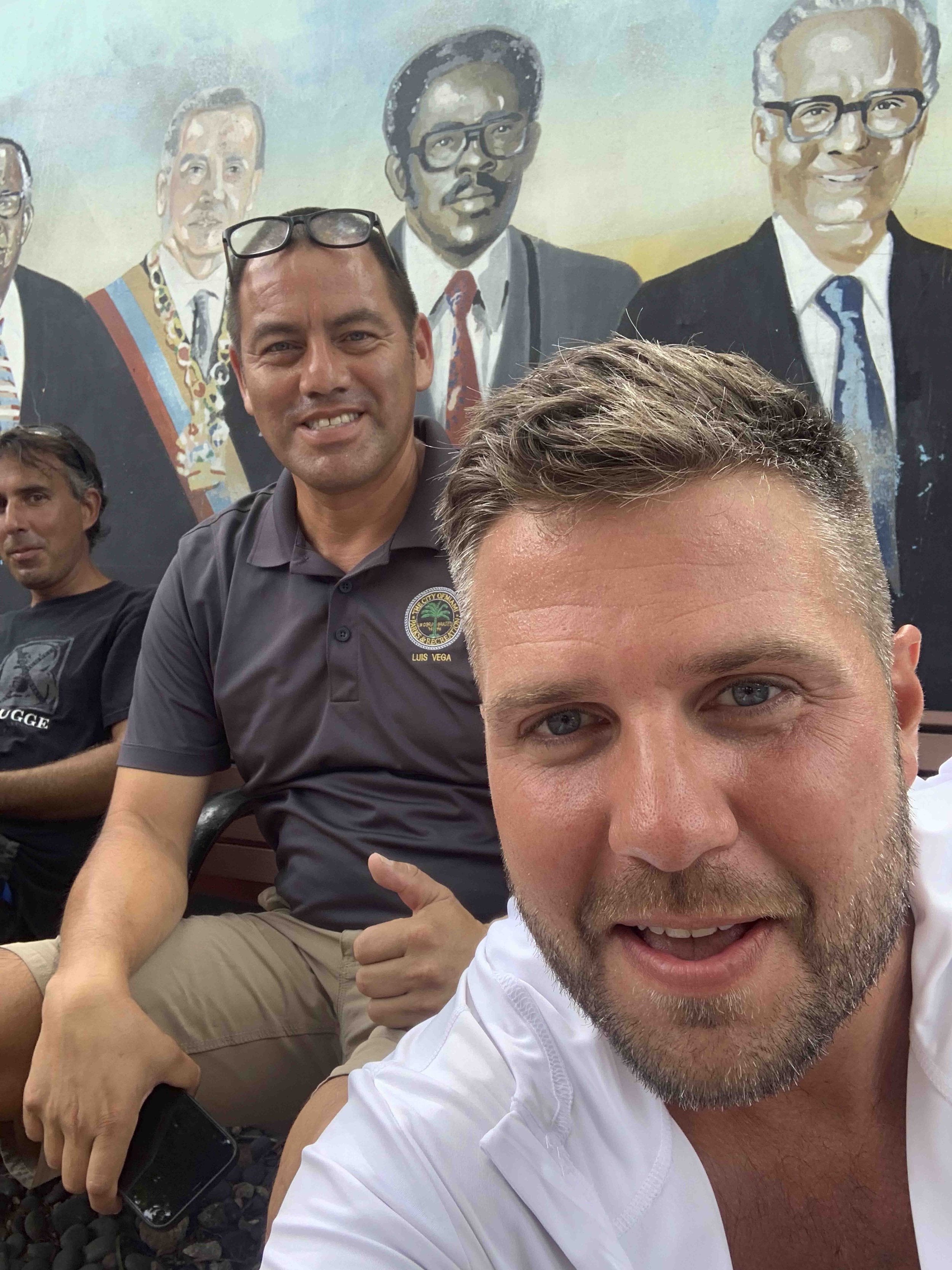 Four men sitting together, with one man in the front taking a selfie. The man in the front has light skin, blue eyes, and short hair, wearing a white shirt. Behind him, there are three other men: one with dark hair, wearing a black shirt, another wit