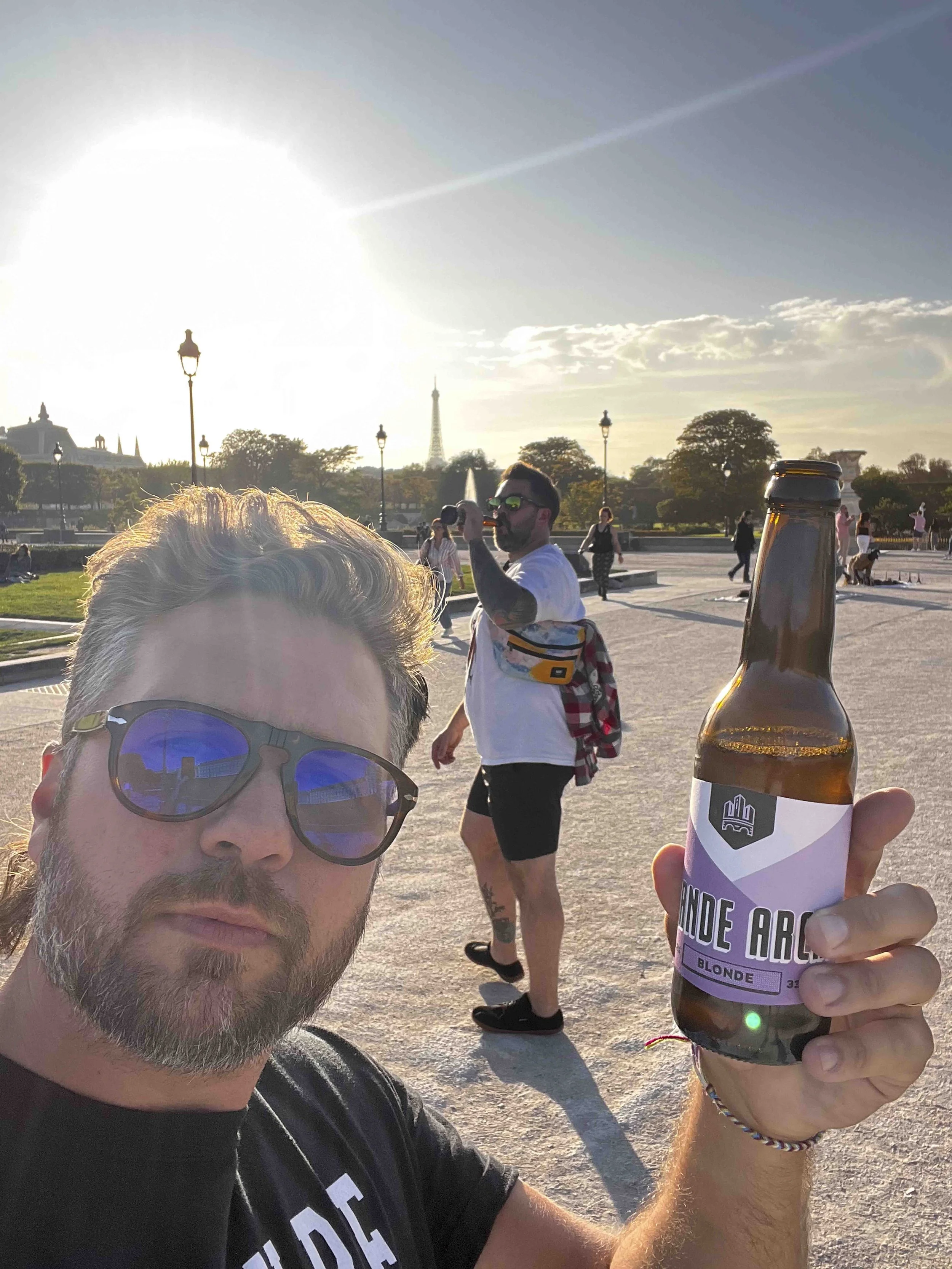 A man with sunglasses and a beard taking a selfie at sunset, holding a bottle of Blonde Ale beer with a purple label. People walk in the background with the Eiffel Tower visible, indicating a Paris park setting.