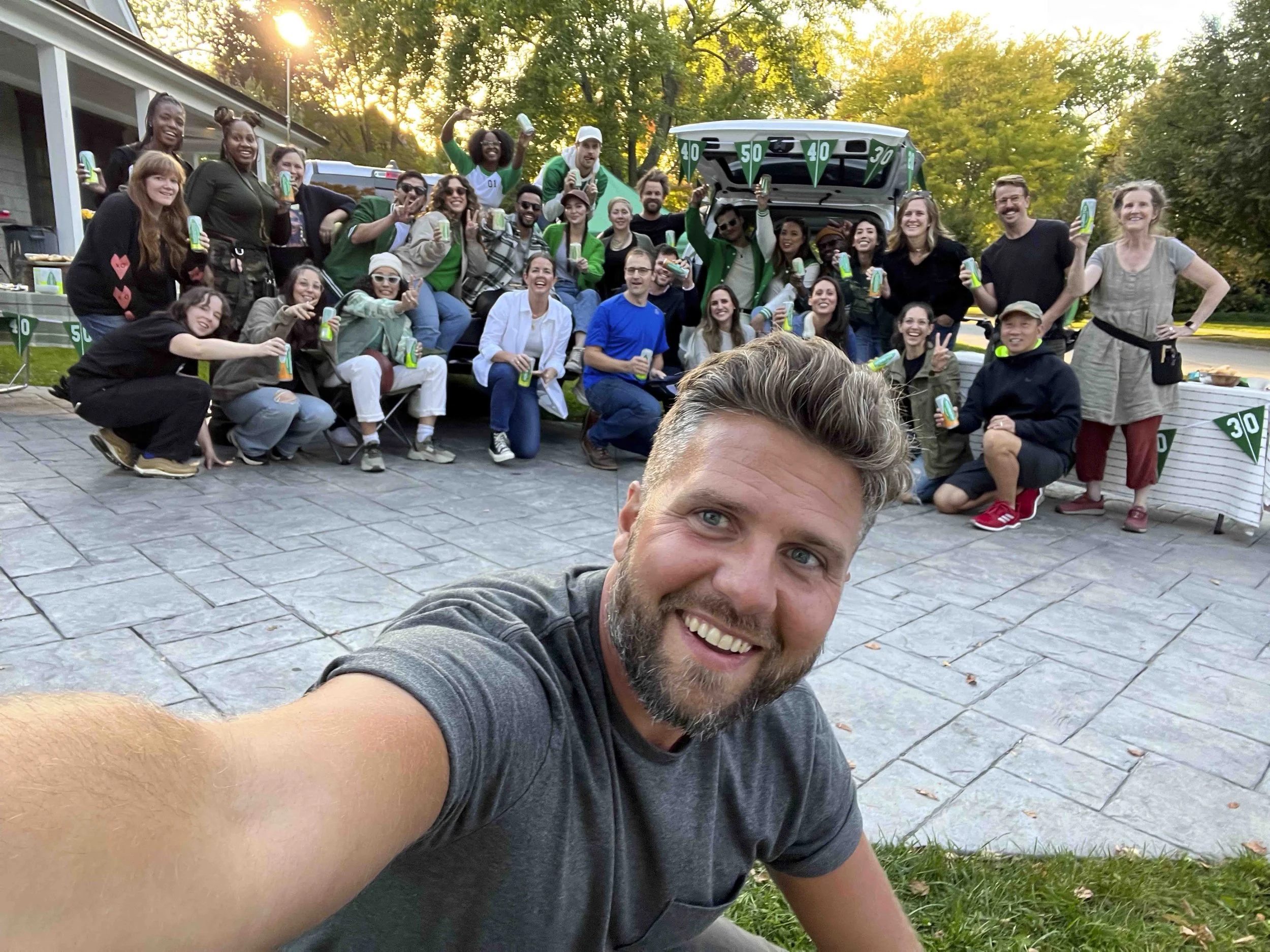 A group of people gathered outdoors during sunset, smiling and holding cans of drinks, with a man taking a selfie in the foreground.