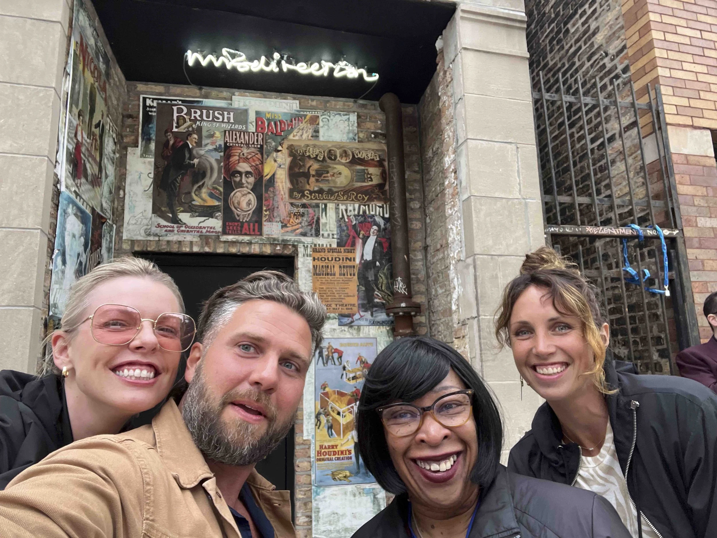 A group of six people taking a selfie in front of posters advertising magic shows and performances, with a brick and stone building in the background.