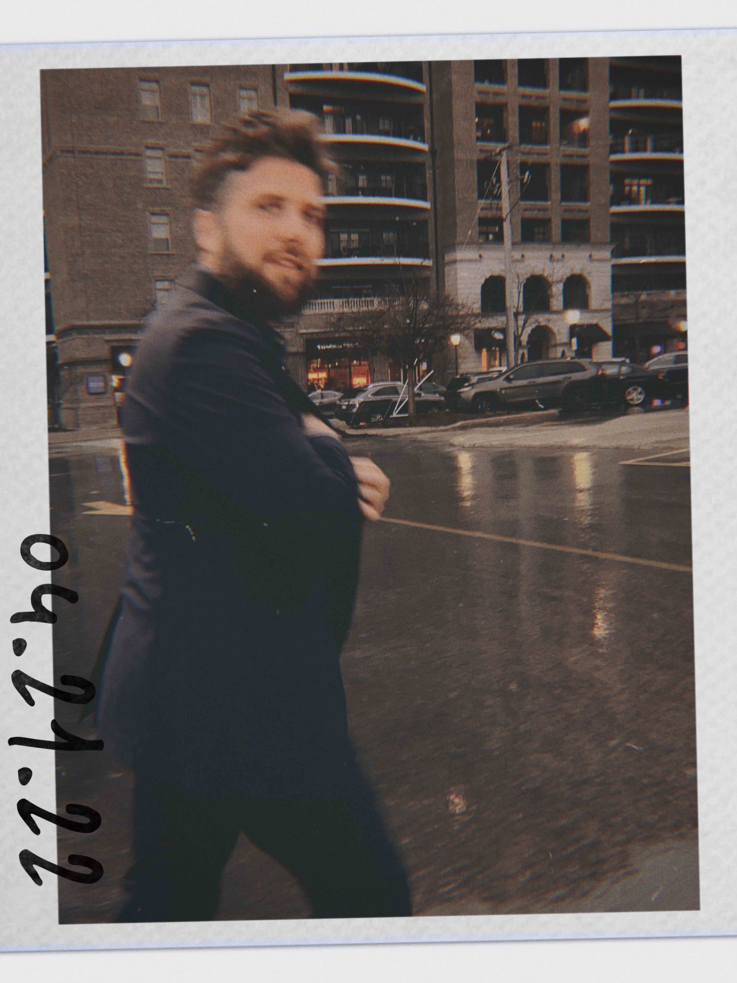 A man with short hair and a beard standing on a wet street at dusk, with a multi-story brick building with rounded balconies and parked cars in the background.