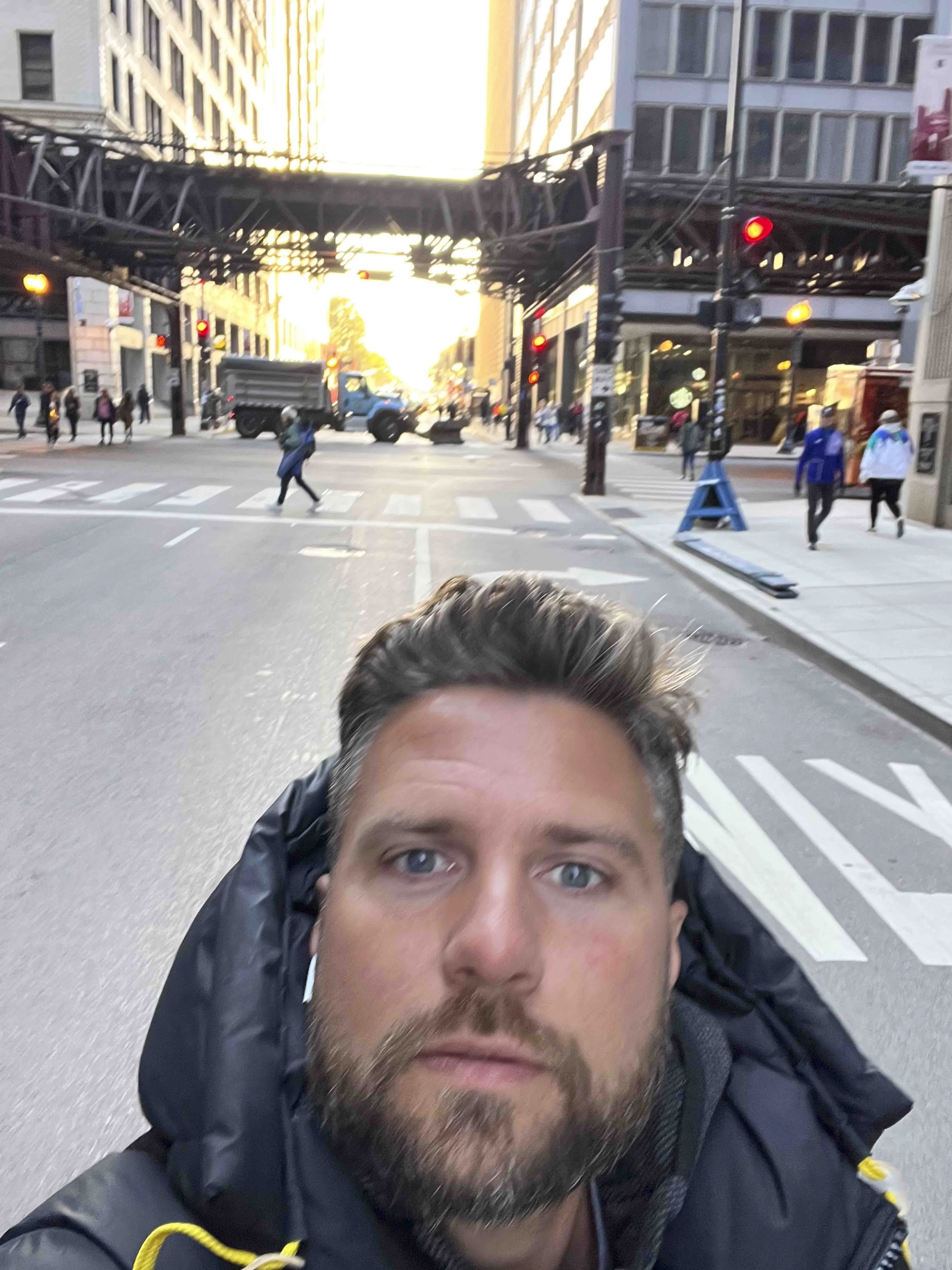 Close-up of a man's face with blue eyes, dark hair, and beard, lying on the ground in a city street with people walking and traffic signals in the background, at sunset.