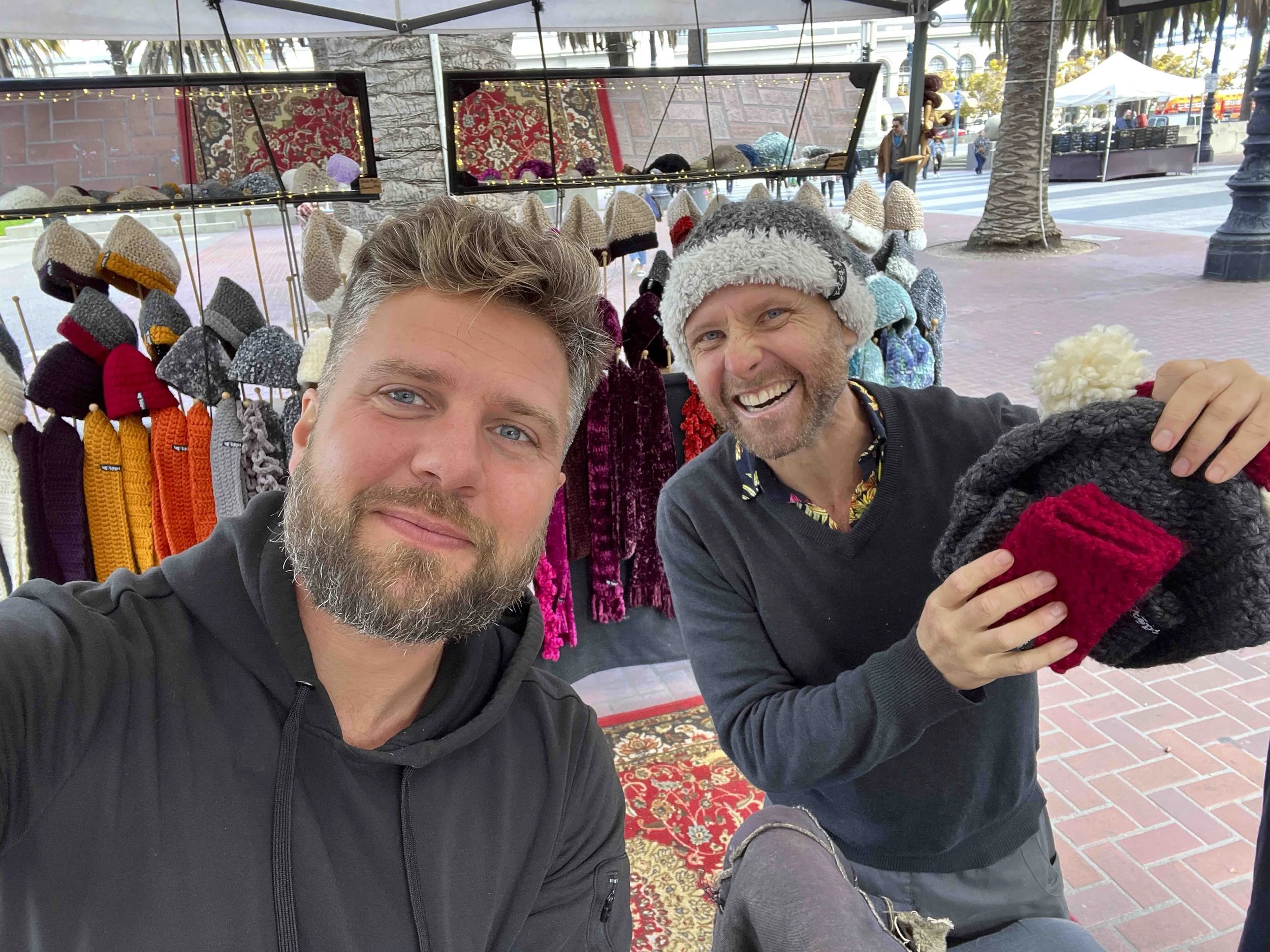 Two men smiling at a street market stall with knitted hats and scarves displayed behind them. One man has light brown hair and a beard, while the other man has light brown hair and is wearing a colorful shirt under a dark sweater.