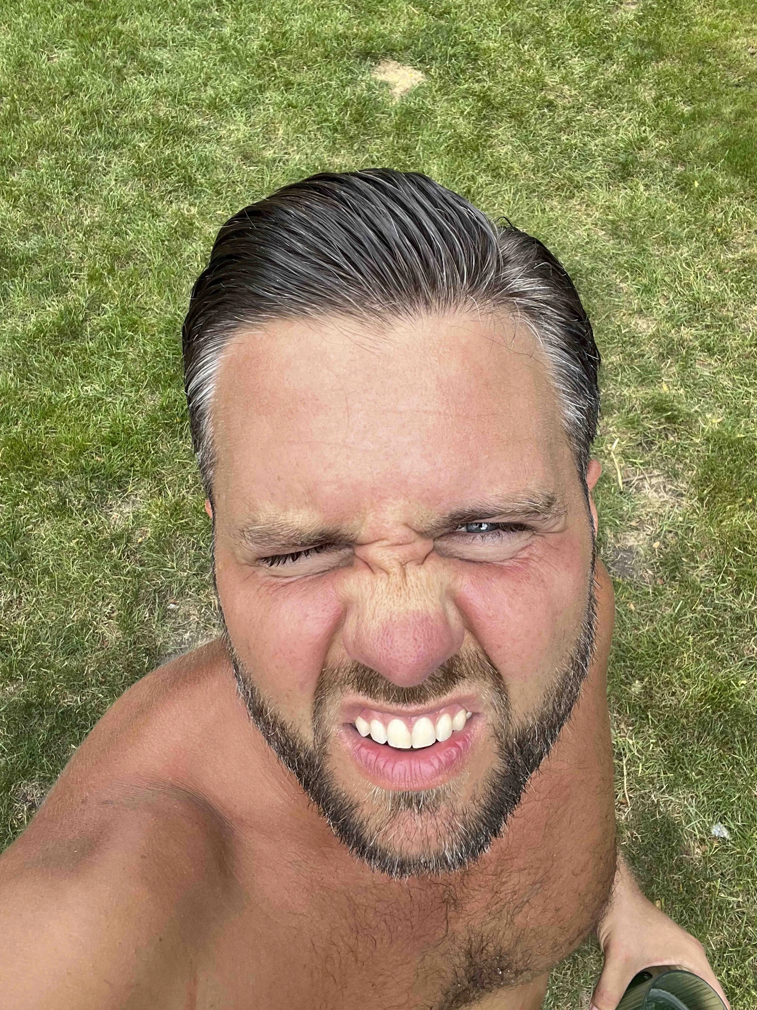 A man with short dark hair and a beard taking a selfie outdoors on grass, showing his face and upper body with a big smile and squinting eyes.