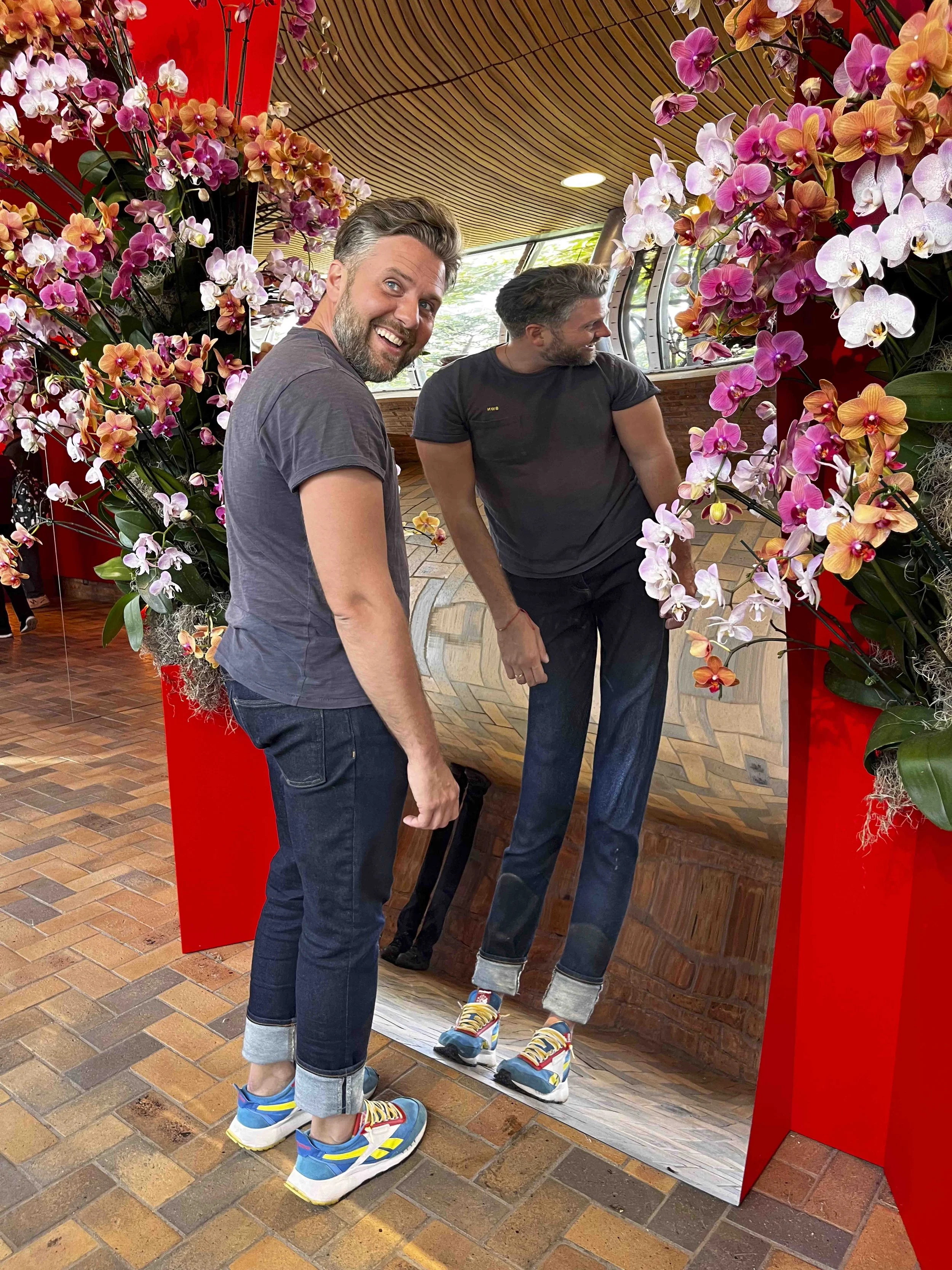 Two men with short hair and beards are posing for a photo behind or inside a mirror framed with pink, orange, and white pansy and orchid flowers. The man on the left is smiling at the camera, wearing a gray t-shirt and jeans rolls. The man on the rig