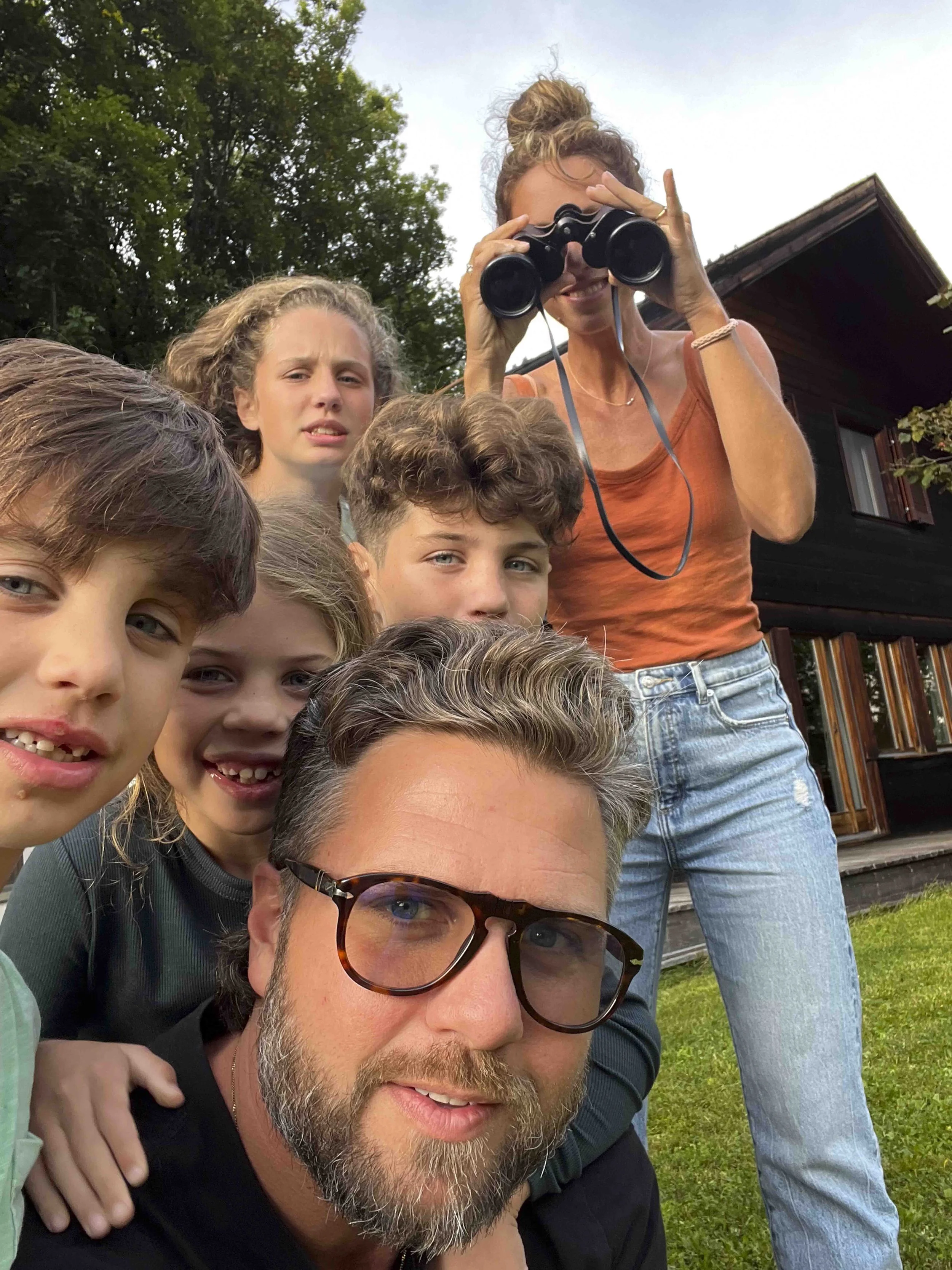 Group of children and an adult man taking a selfie outdoors, with some children and a woman in the background, wearing sunglasses and casual clothing, in front of a house with a wooden exterior.