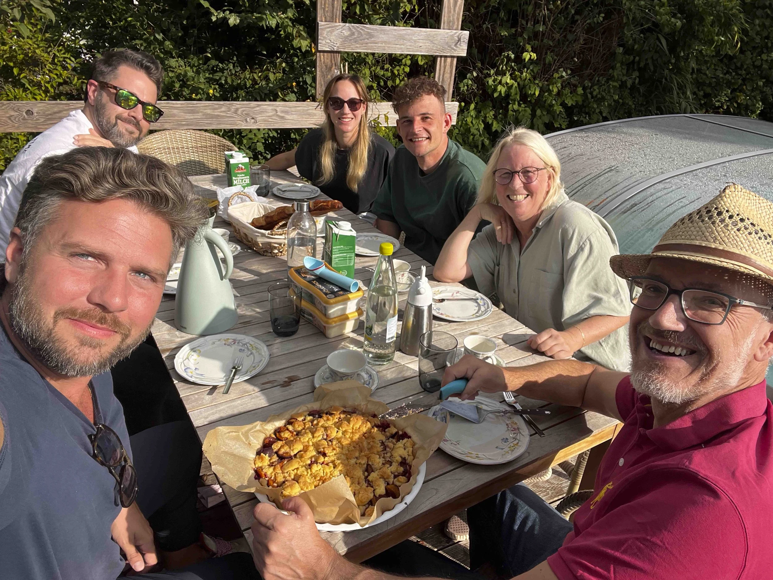 Group of six people sitting around a wooden outdoor table, enjoying a meal, with a large flat cake in the foreground, drinks, and a carton of milk, in a garden setting with green foliage.