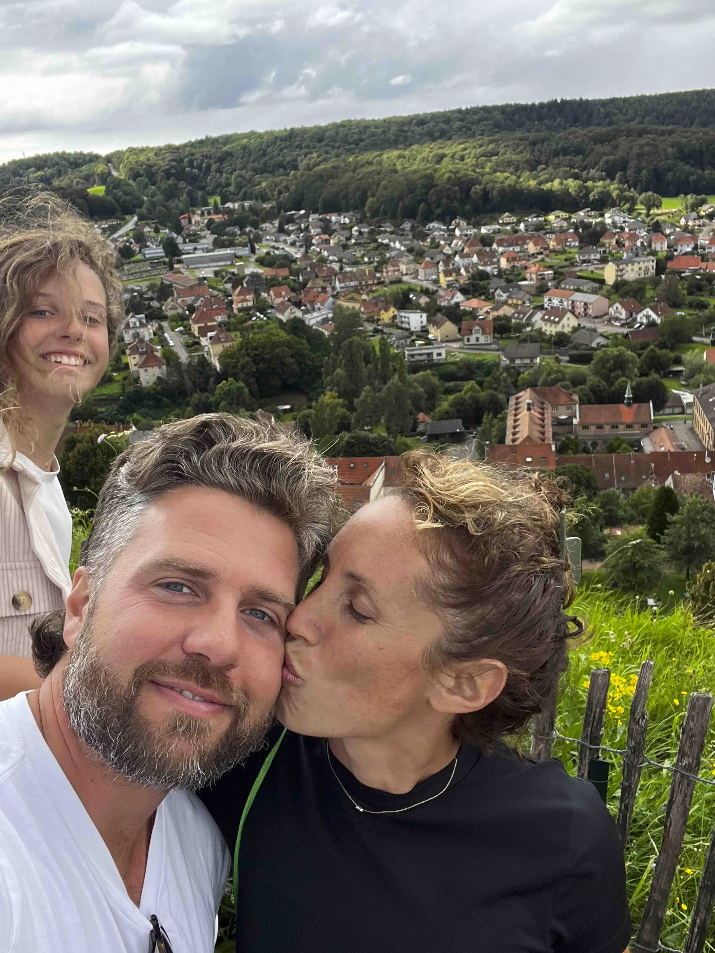 A group of people taking a selfie outdoors with a hillside town and green forested landscape in the background. Two women are visible, one is smiling at the camera, and the other is kissing a man on the cheek. The scene is overcast.