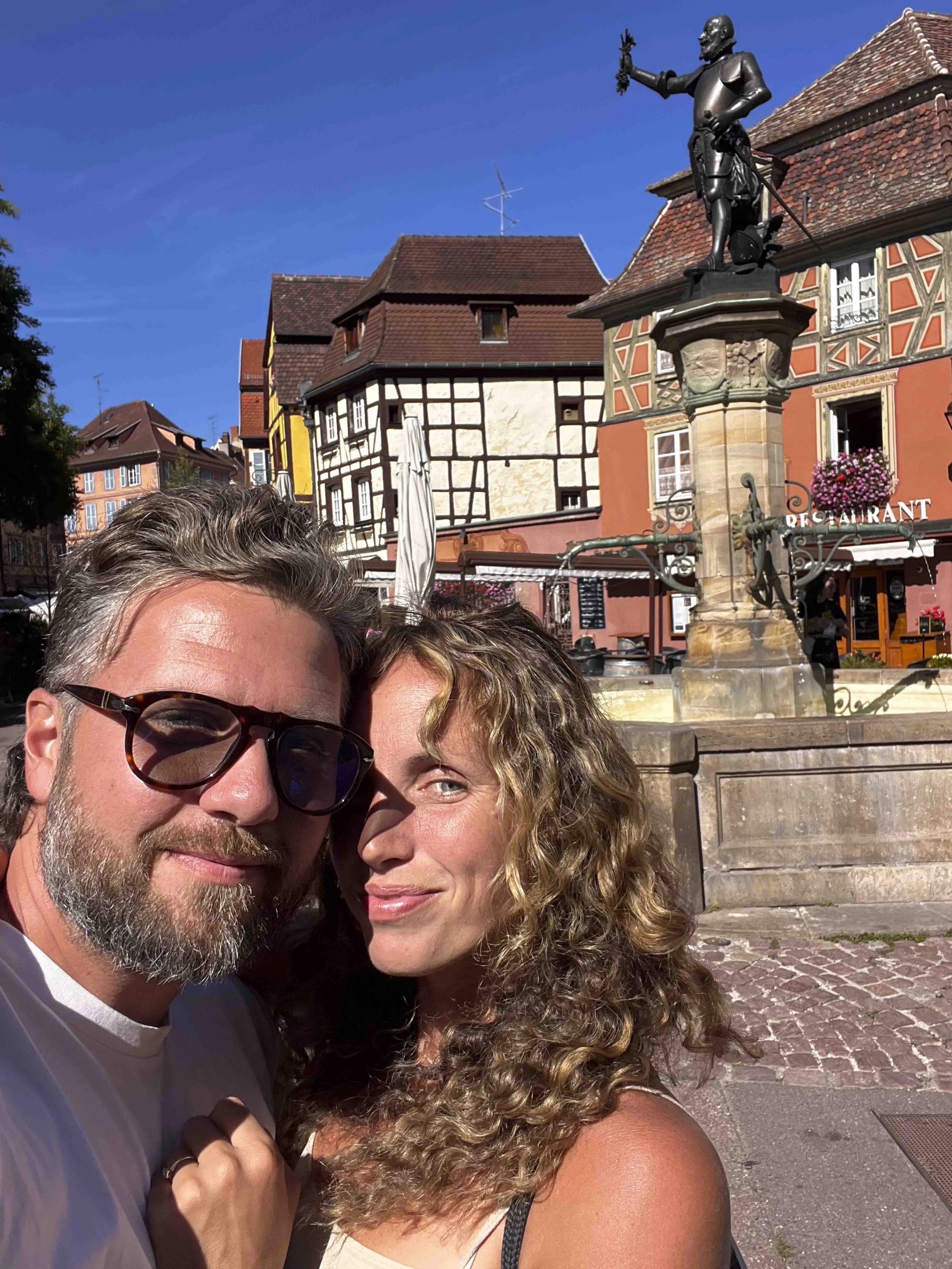 Close-up of a man and woman smiling with a European town square in the background, featuring traditional architecture, a statue, and a restaurant.