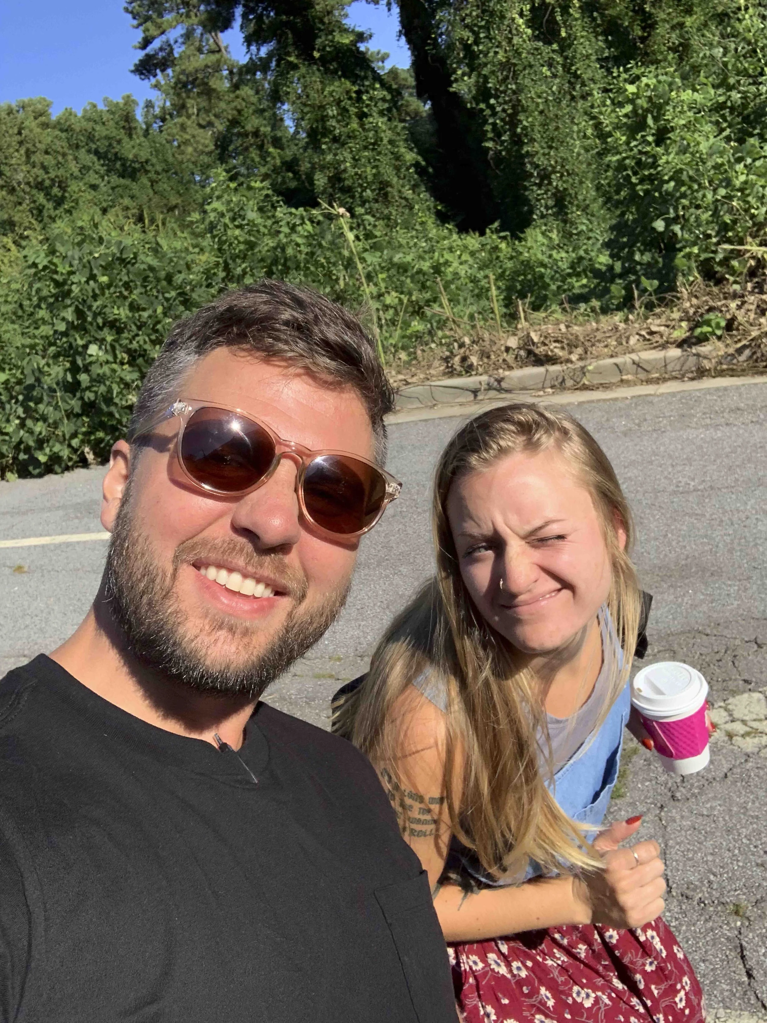 A man with short brown hair, a beard, and wearing sunglasses taking a selfie, with a young woman with long blonde hair, squinting and holding a cup of coffee, sitting on a curb on a sunny day with greenery in the background.