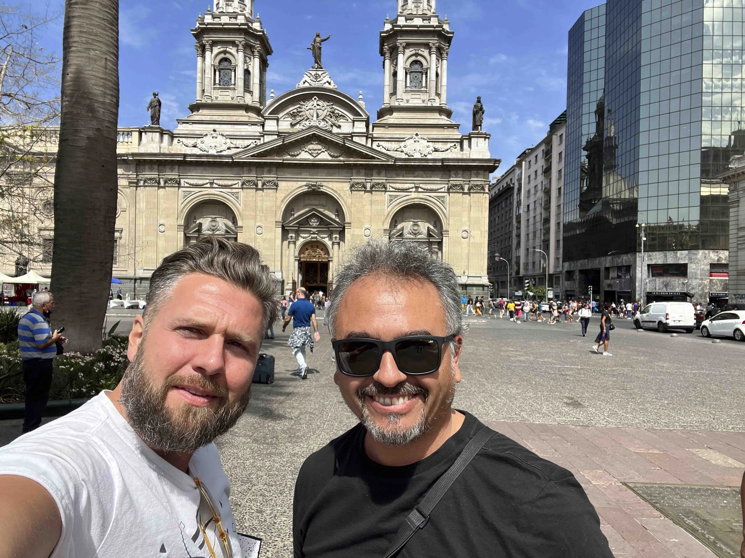 Two men taking a selfie in front of a historic church with classical architecture, large arched entrance, and statues on the facade, with a modern glass building nearby, during a sunny day with people walking in the background.