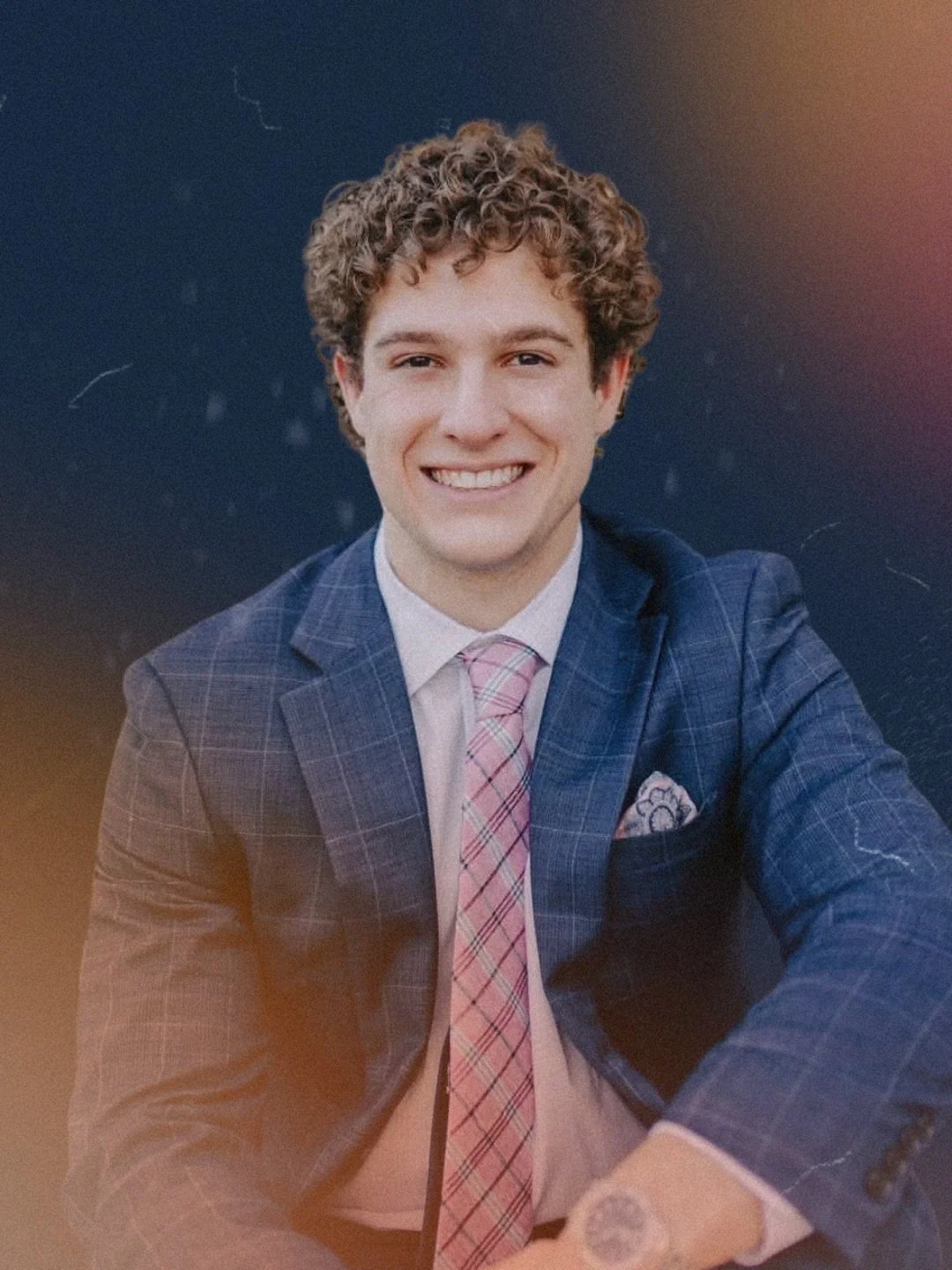 Portrait of a young man with curly hair wearing a blue checkered suit, pink checkered tie, and white shirt, smiling at the camera.