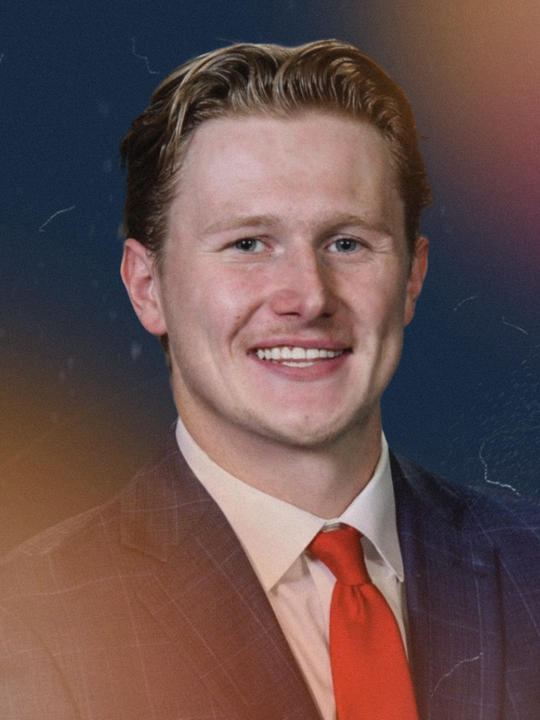 A young man with short, wavy red hair, wearing a dark plaid suit, a white dress shirt, and an orange tie, smiling at the camera against a dark, abstract background.