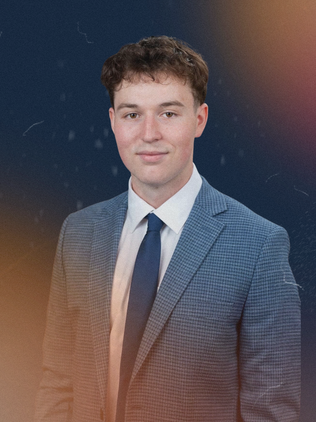 A young man with short brown hair wearing a blue checkered suit, white shirt, and dark blue tie posing against a dark, abstract background with soft light.