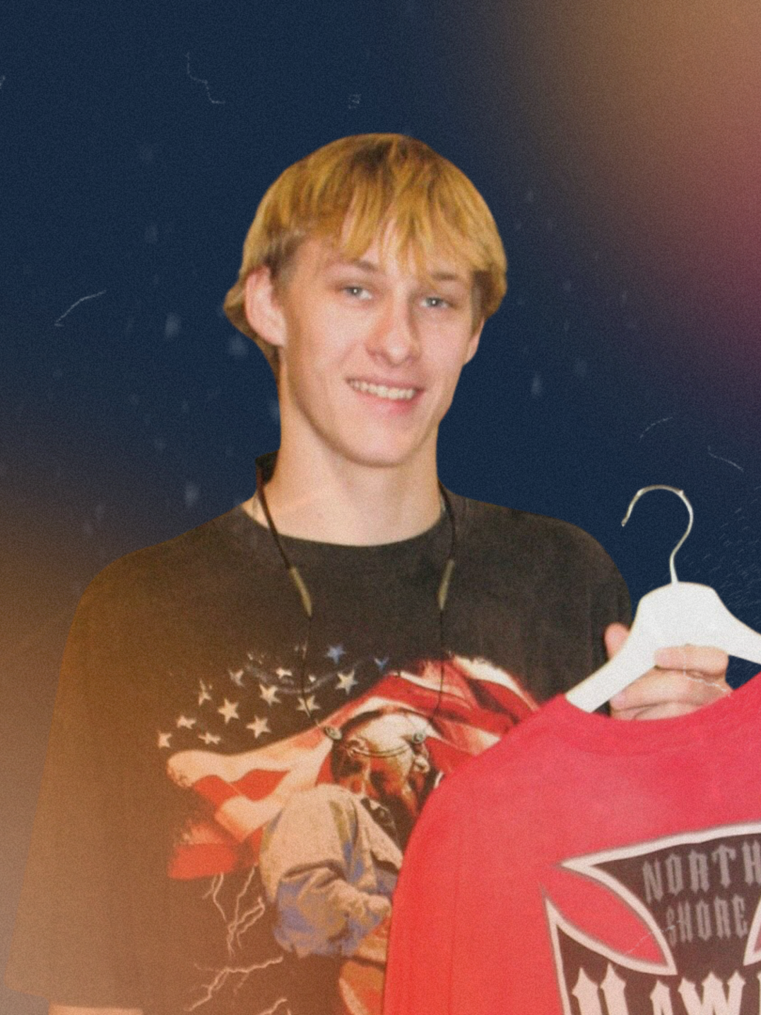 A young man with blond hair smiling, wearing a shirt with an American flag design, holding a white hanger, standing in front of a dark background with light streaks.