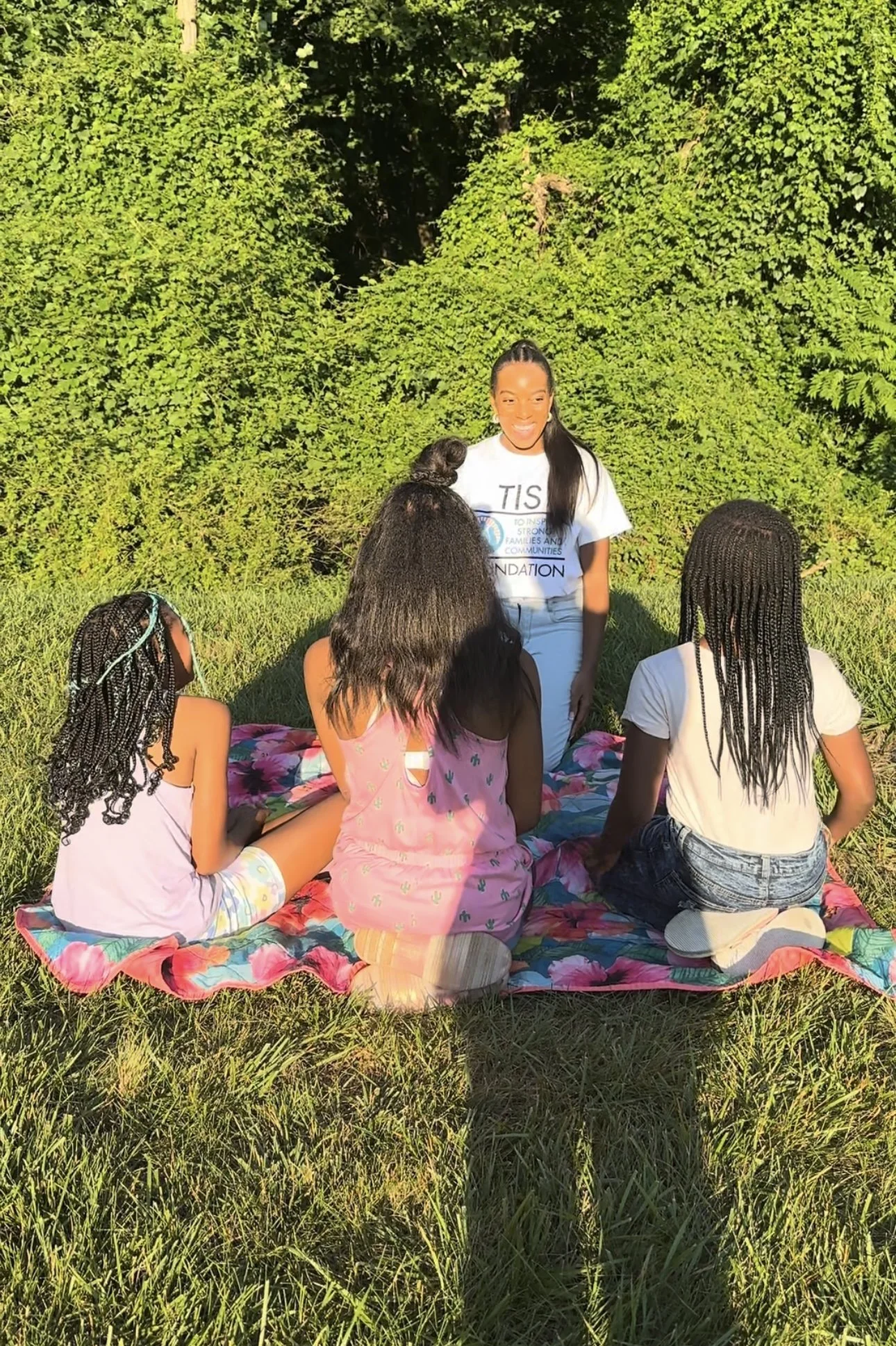A woman standing in front of four girls sitting on a floral blanket outdoors with green trees in the background.