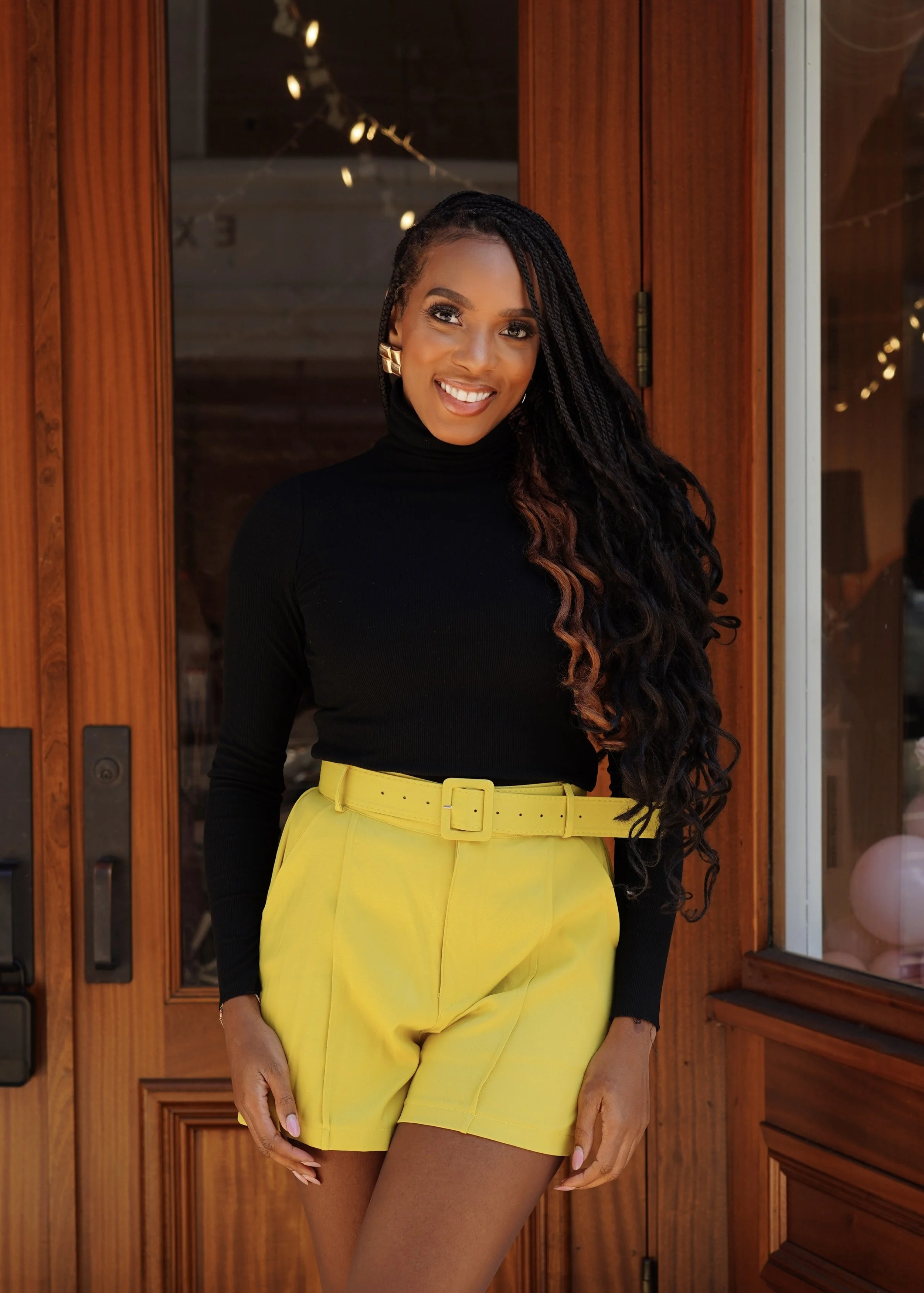 Smiling woman in black turtleneck and yellow shorts standing in front of a wooden door.