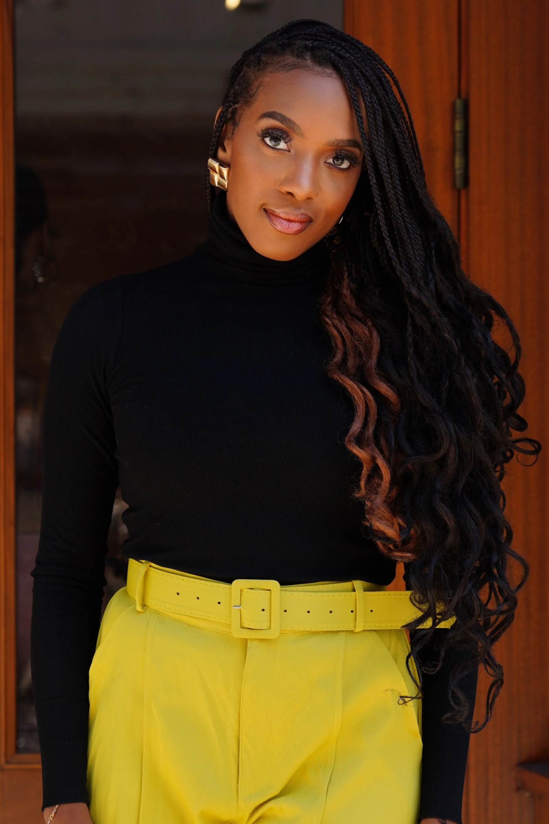 A woman with long, wavy black hair and gold earrings standing in front of a wooden door, wearing a black turtleneck and bright yellow pants with a matching belt.