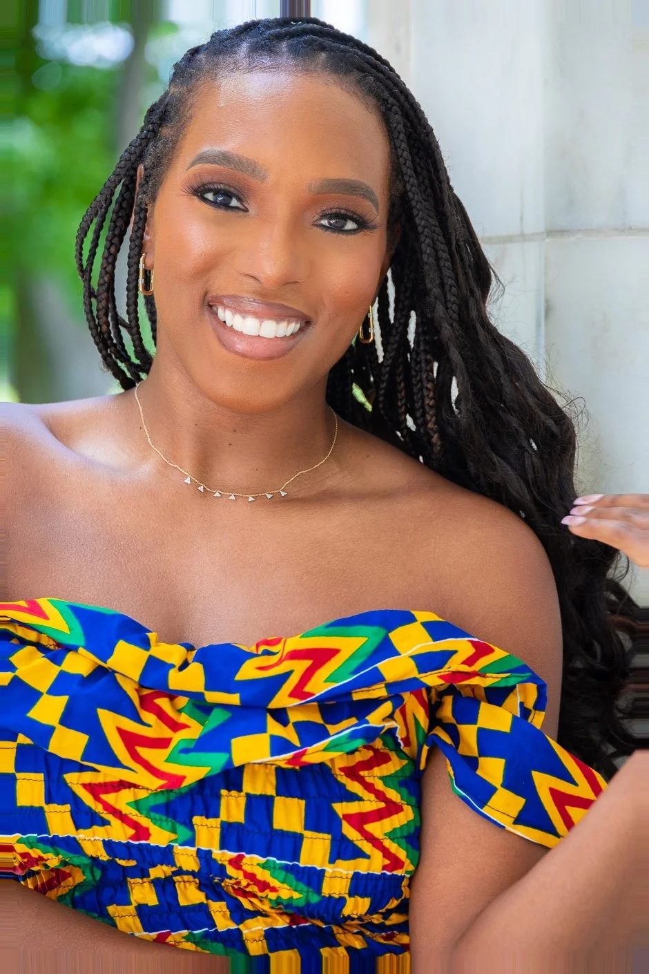 A smiling woman with dark braided hair, gold hoop earrings, and a delicate necklace, wearing a colorful, off-the-shoulder dress with a vibrant, geometric pattern, posing outdoors.