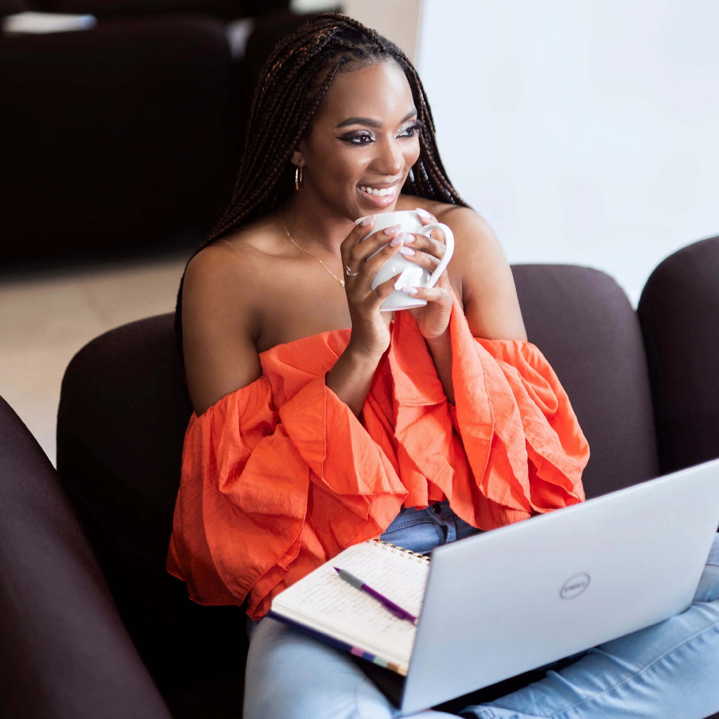 A woman with braided hair, smiling, sitting on a couch with a laptop, a notebook, and a pen, holding a mug and wearing an off-the-shoulder orange top.