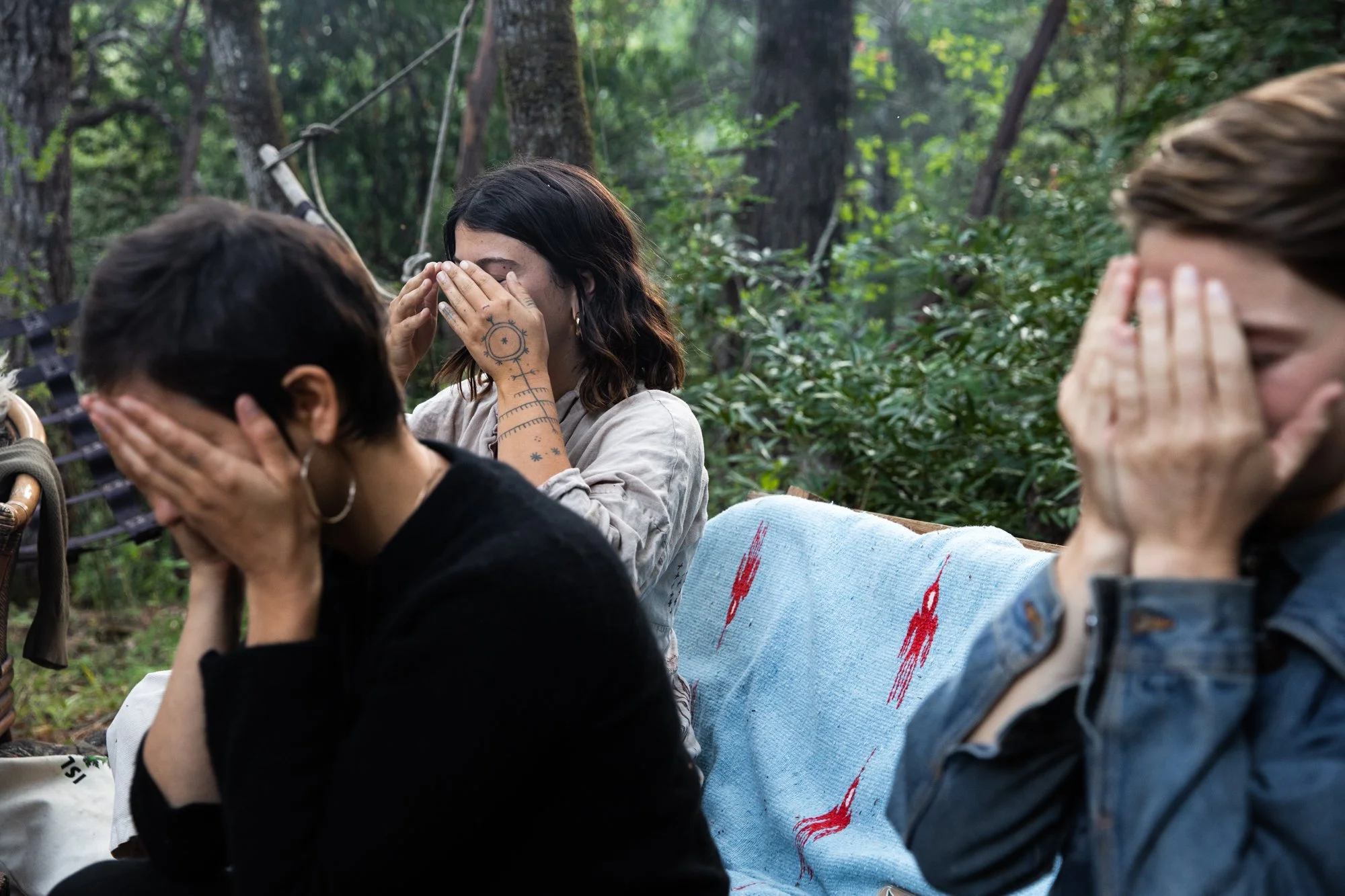 Image from PRAXIS, a photo essay shot in California by photographer Kaare Iverson. Three women sitting outdoors in a wooded area, covering their faces with their hands, possibly in prayer or reflection.
