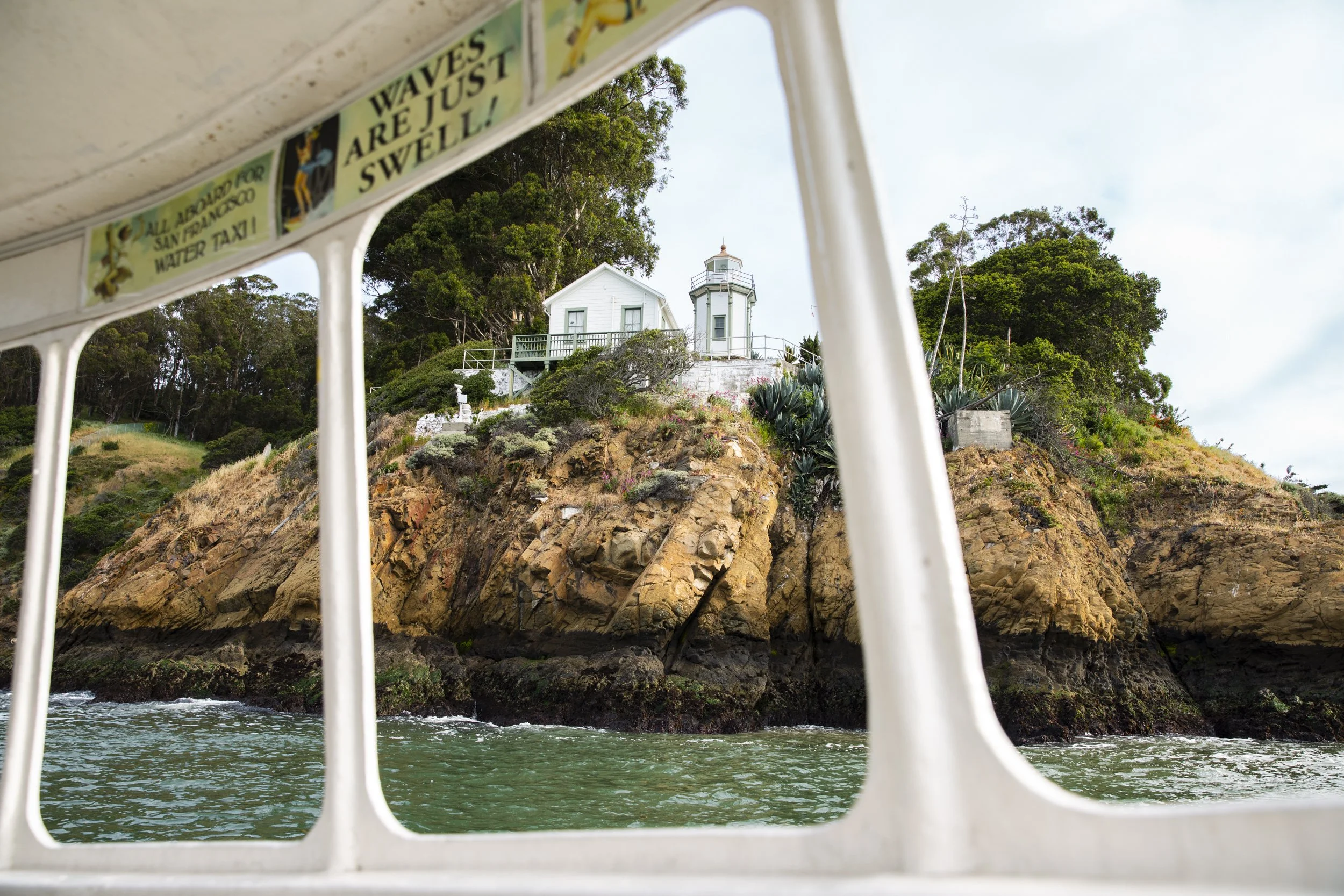 POV view through a water taxi window of a rocky coastline with a white lighthouse surrounded by trees.  Shot by California-based commercial photographer Kaare Iverson for Treasure Island Development Group's lifestyle advertising campaigns.