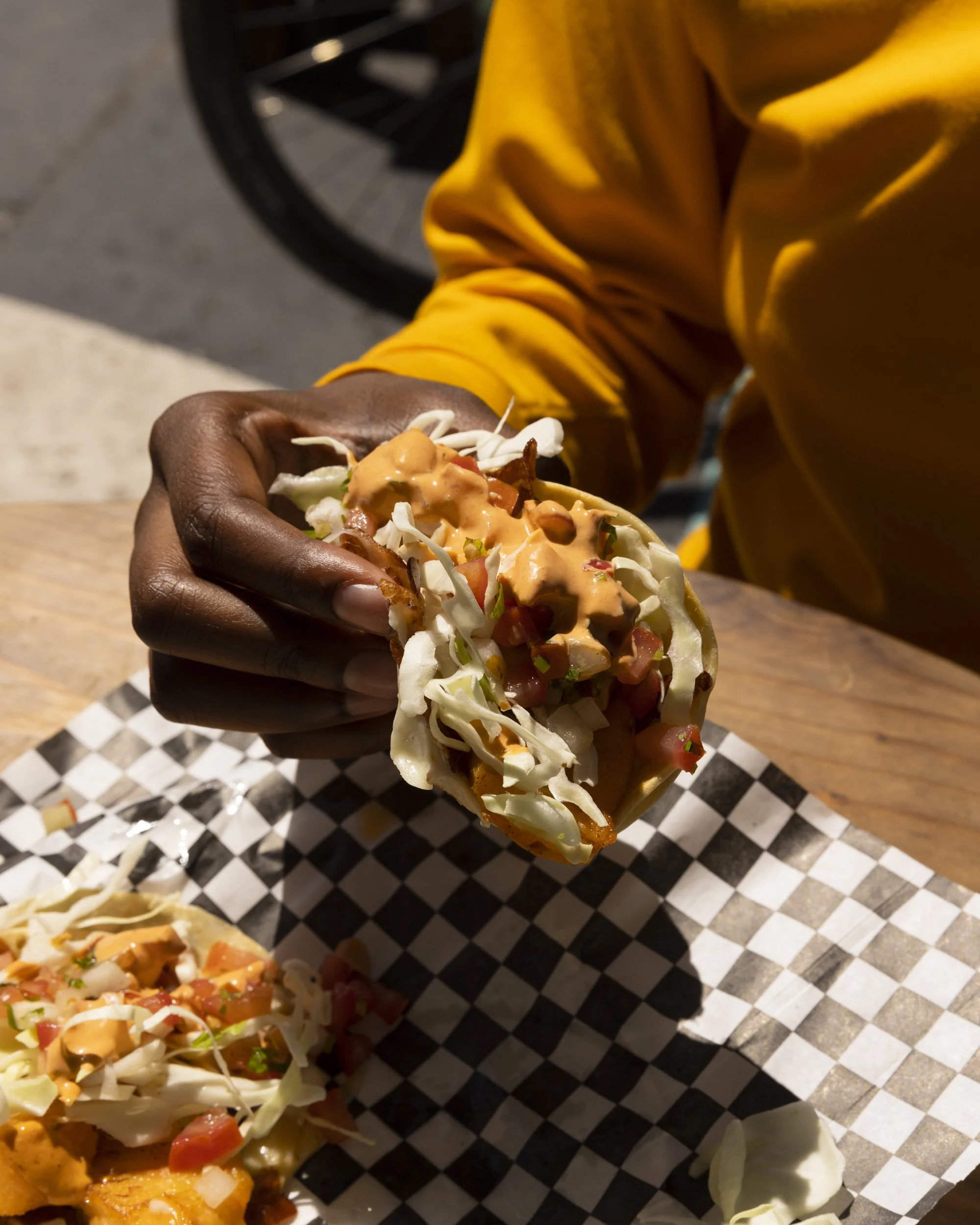 Food adventure lifestyle travel photography by commercial photographer Kaare Iverson in California.  A hand holding a taco with shredded lettuce, diced tomatoes, shredded cheese, and sauce, on a checkered paper-lined tray.