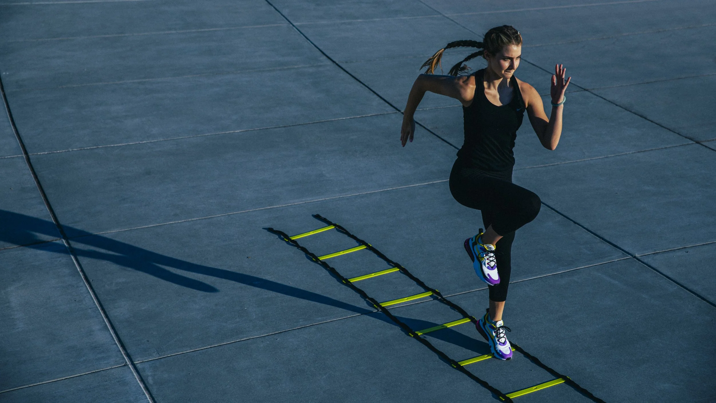 Commercial fitness photography for a mobile application of a woman dressed in black workout clothes and running shoes performs cardio exercises on an urban paved area, using a small agility ladder - shot in California.