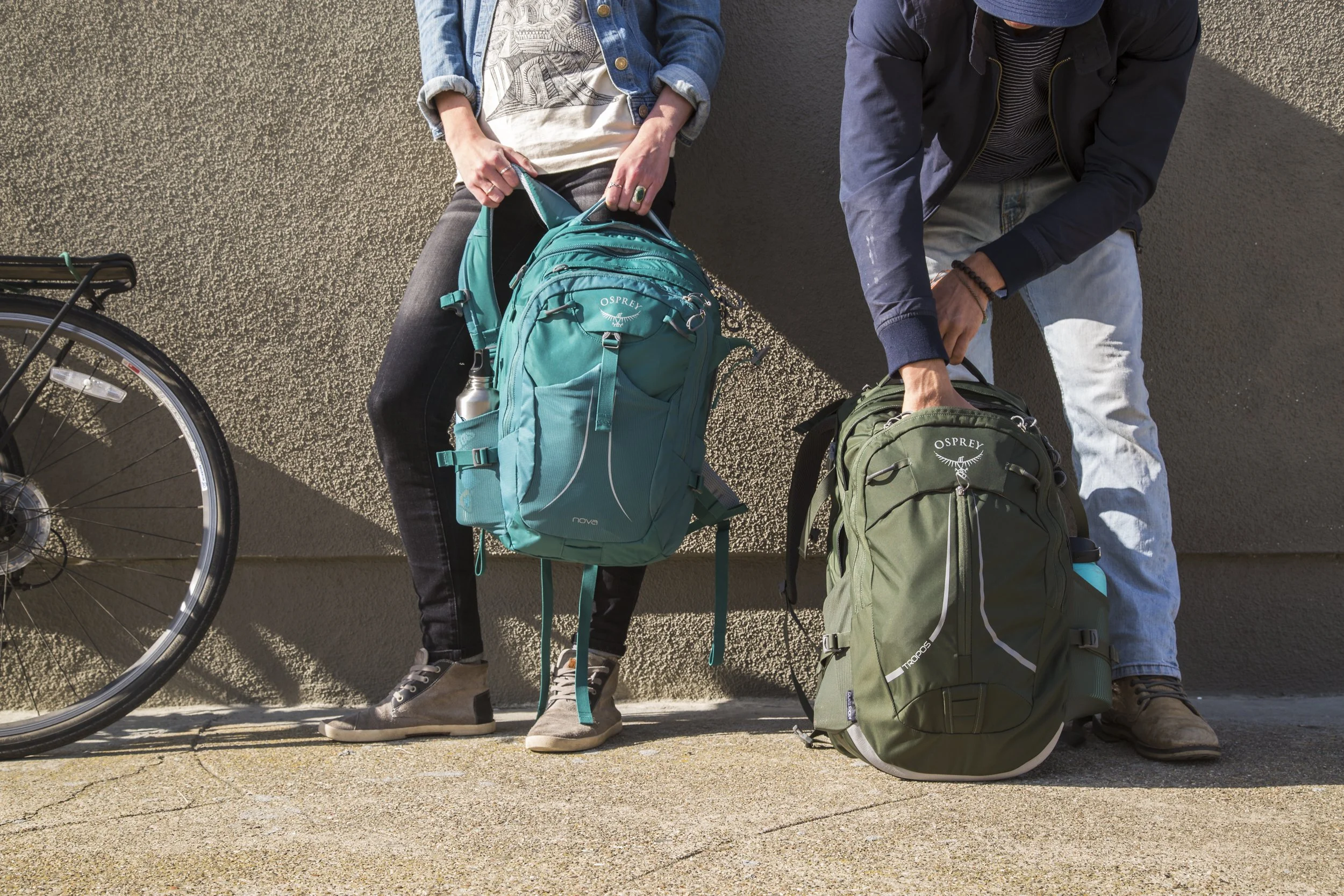 Two people with Osprey backpacks standing against a wall, with part of a bicycle wheel visible on the left side, and a shadow cast on the wall. Shot by California commercial photographer Kaare Iverson.