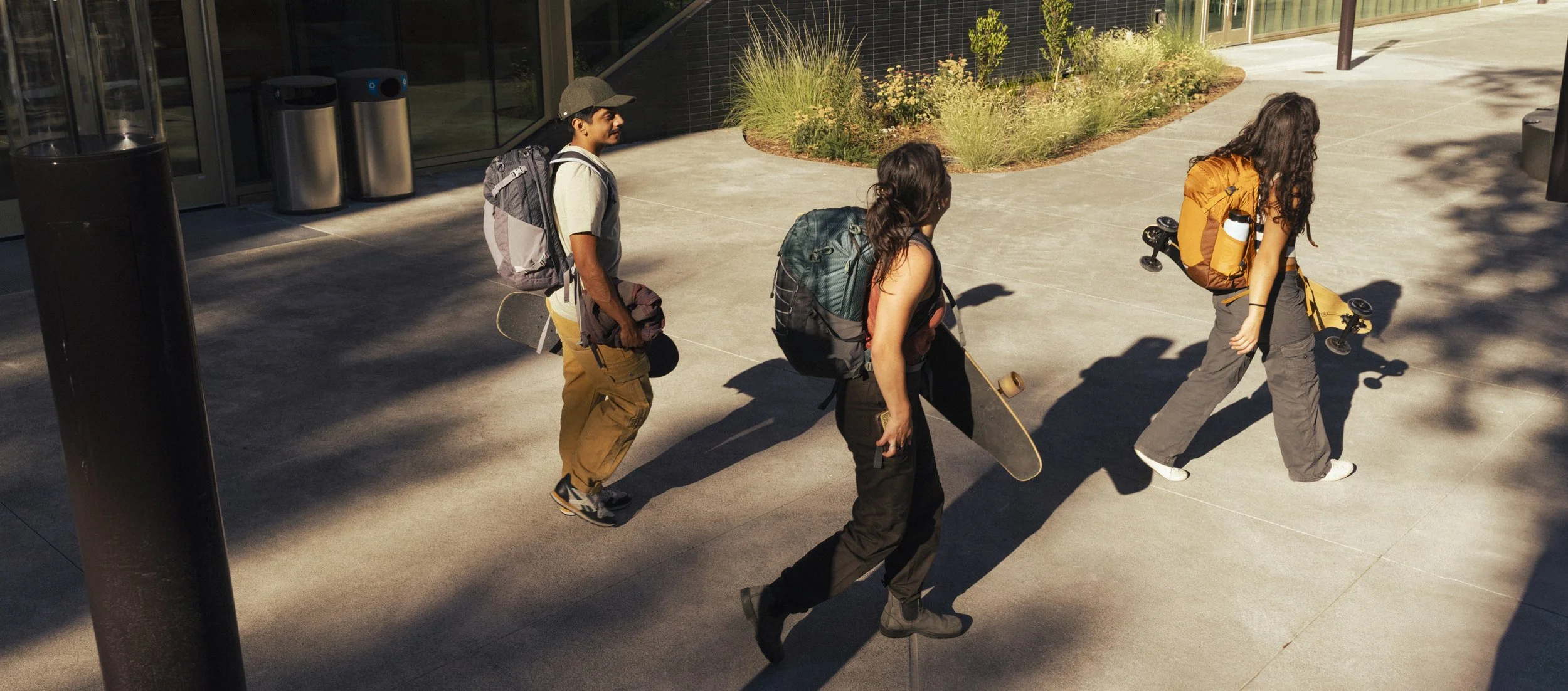 Three young people walking with skateboards and Osprey backpacks outside a modern college building on a sunny day. Shot by California commercial photographer Kaare Iverson in Sonoma County.