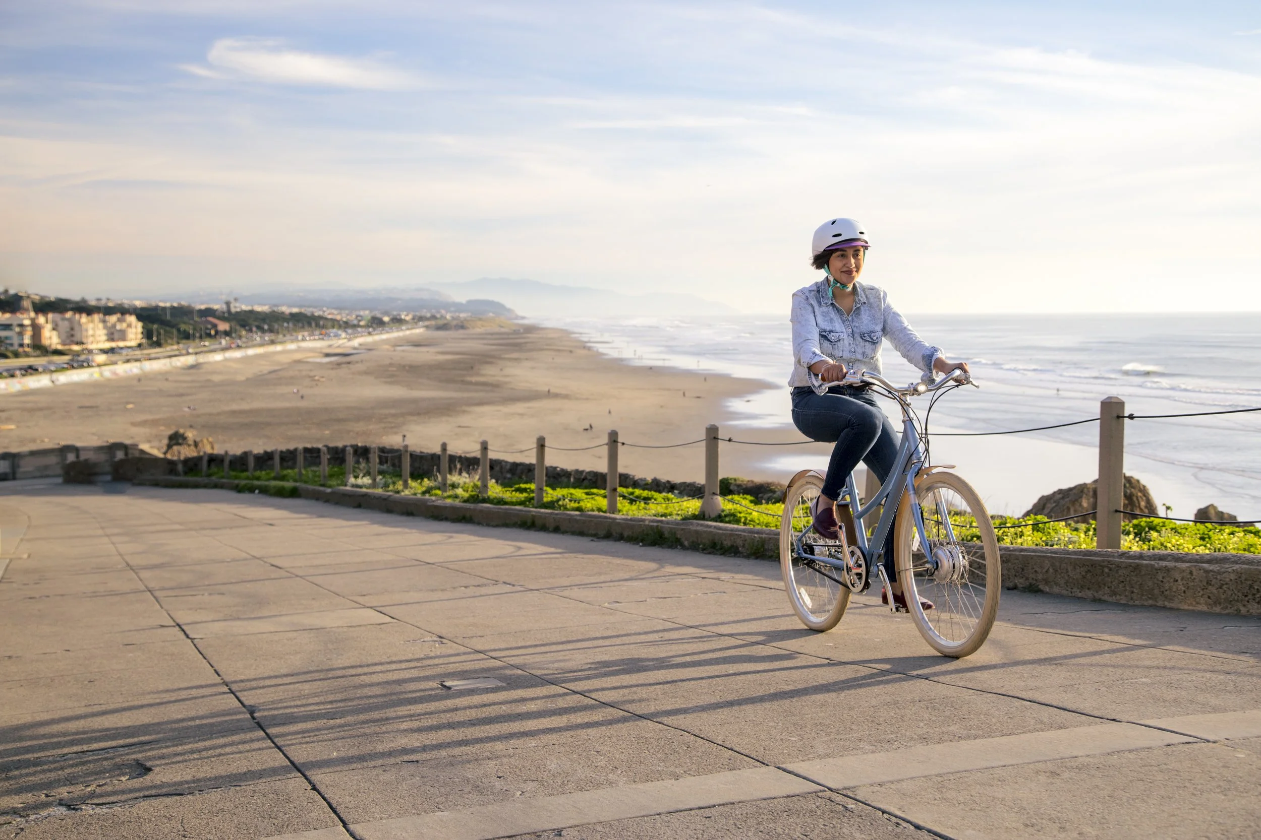 A woman riding a e-bicycle along a coastal promenade at sunset, with a sandy beach and ocean on one side and buildings on the other. Shot by California commercial photographer Kaare Iverson.