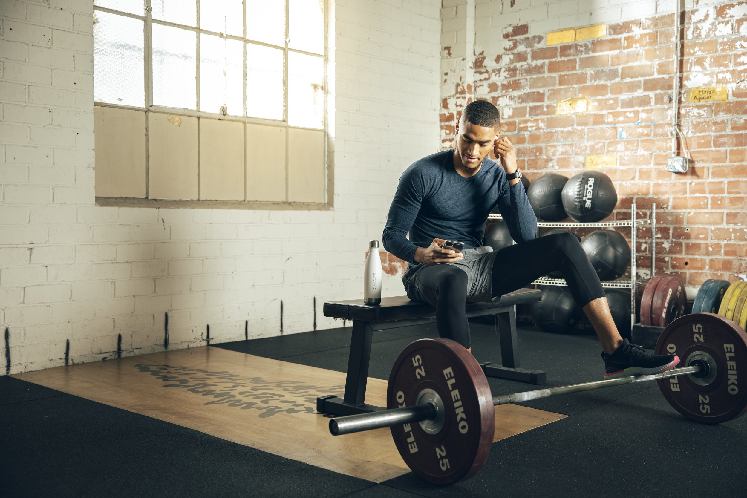 Athletic fitness and training photography by Bay Area commercial photographer Kaare Iverson for Future Fit App. A young man with short hair and gymwear reviews his workout on his phone at a bench in a crossfit gym.