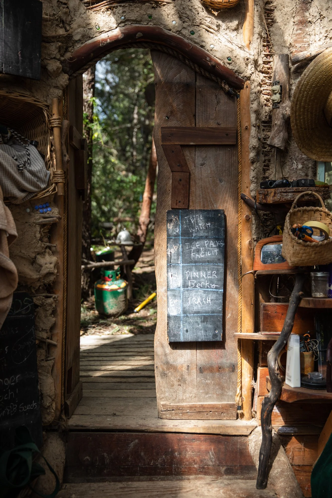 Image from the photo essay PRAXIS about alternative living in California by photographer, Kaare Iverson. Wooden door with a chalkboard sign listing items to do and trash bin outside in a rustic, outdoor setting.