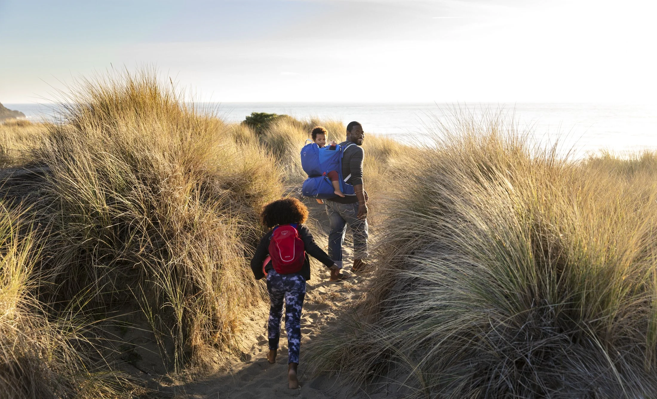 Product lifestyle photography on the California coast by commercial photographer Kaare Iverson. A family of three walking through sand dunes covered with tall grass, near the coast for Osprey Packs product line.