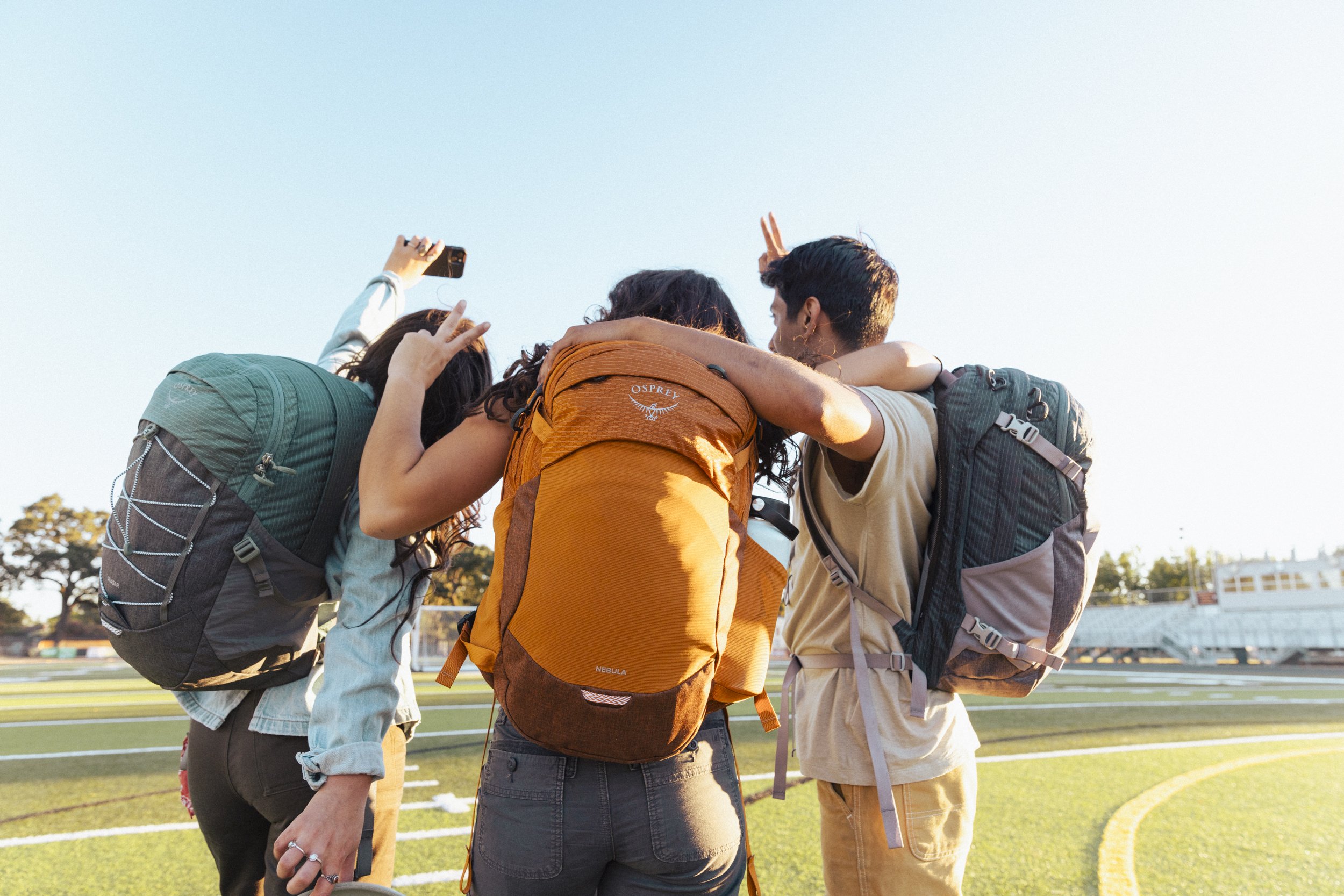 College lifestyle photography by commercial photographer Kaare Iverson for Osprey Packs. Three friends with backpacks taking a selfie together on a sports field during sunset.